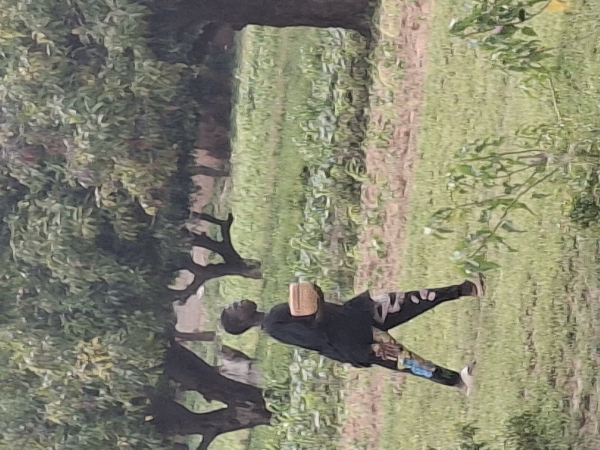 In Kwadon, Nigeria, a young boy stands in a grassy field with scattered trees. He is wearing a black long-sleeved shirt and patterned pants. In his hands, he holds a light brown, shallow bowl. The boy is looking towards the right of the frame. The background consists of more trees and dry, reddish-brown earth. The overall lighting suggests an overcast or early morning/late afternoon setting.