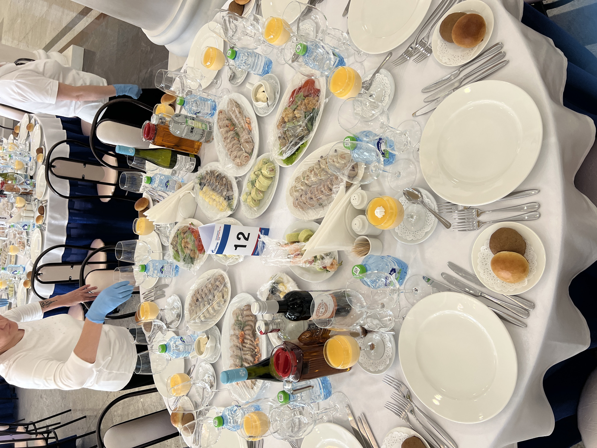 This overhead image depicts a long, formally set table for a banquet or corporate event, likely indoors in Votkinsk, Russia. The table is covered with white tablecloths and features multiple place settings, each with a large white oval plate, a full array of silverware, and various glasses for water, wine, and orange juice.
Numerous appetizers and cold dishes are arranged along the table in white oval platters, all covered with clear plastic wrap, indicating they are awaiting service or protecting their contents. These dishes appear to contain sliced meats, possibly rolled preparations, and salads. Small plates holding two types of bread rolls are placed at each setting. Bottled water and other beverages, including red wine and a clear spirit, are also distributed across the table.
Catering staff, dressed in white uniforms and blue gloves, are visible in the scene, actively engaged in setting up or attending to the table, suggesting preparations are underway for guests. A prominent white table number card displays the number "12" and features the logo and text "ГАЗПРОМ СОЦПРОФ," indicating a connection to Gazprom, a Russian company, likely for a corporate social event or a trade union gathering. The scene conveys an atmosphere of organized readiness for a professional event.