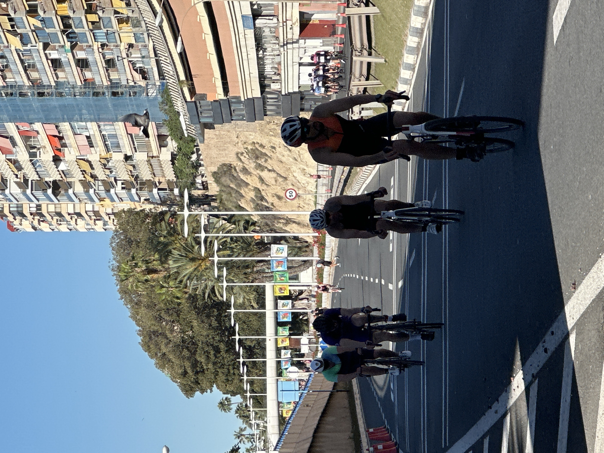 The image shows several cyclists riding in a race or event on an urban road. The location appears to be a coastal city based on the presence of the sea, urban development, and palm trees. Several cyclists are visible in the foreground, with a larger group further ahead. The lead cyclist wears an orange top. Buildings line the road, including a high-rise apartment complex with colorful awnings. There are decorations along the roadside, possibly banners or flags, that could be associated with the event. A speed limit sign with the number "50" is also present. The weather seems sunny and clear.