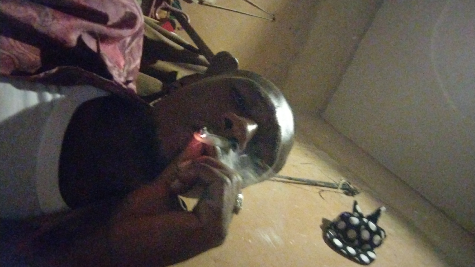 The image, captured in Jos, Nigeria, depicts an individual engaged in smoking an unknown substance from a pipe in a dimly lit indoor environment. The subject's head, right hand, and part of their torso are visible, with their skin appearing dark. They are holding a dark, possibly wooden or calabash pipe to their mouth, while simultaneously using an ignited lighter in their right hand to apply flame to the pipe. Wisps of smoke or vapor are seen rising from the pipe, indicating the act of lighting and inhaling.
The background consists of a plain, light-colored wall and floor, suggesting a simple room. On the lower left, draped fabric with a purple and red pattern is partially visible. A small, black object adorned with white polka dots, possibly a decorative item or a miniature hat, rests on the floor in the lower right. The low light conditions suggest the photograph was taken at night or in a poorly illuminated room. No visible text is present.