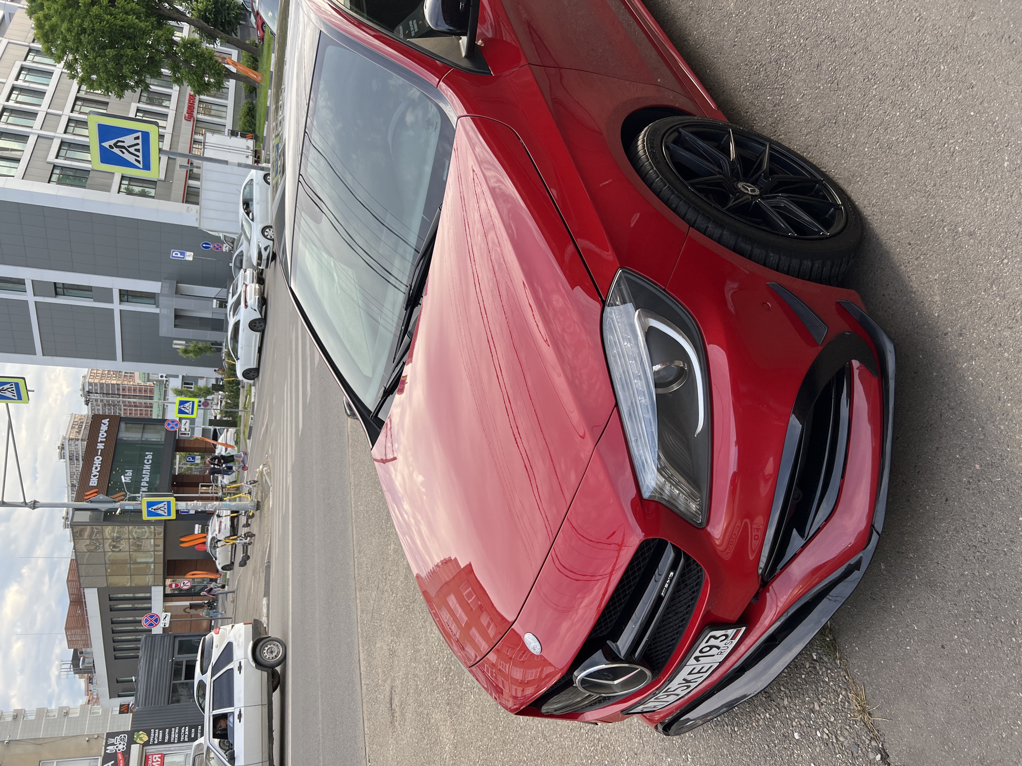 This media file captures an urban street scene in Krasnodar, Russia, under bright daylight. The main subject is a glossy red Mercedes-Benz car, likely an A-Class or CLA-Class, prominently featured on the right side of the road. Its front-right quarter is visible, showcasing black multi-spoke wheels, the Mercedes-Benz emblem on the grille, and "AMG" branding. The Russian license plate "T795KE 193 RUS" is clearly displayed, with "193" being the region code for Krasnodar Krai.
The setting is a bustling street lined with multi-story buildings, some appearing commercial with large windows, and others residential. Reflections on the car's surface indicate clear, sunny weather. Everyday urban activity is taking place, with other vehicles (mostly white cars) parked along the curb and driving on the road in the background. Pedestrians can be seen walking on the sidewalks, and a row of yellow electric scooters is parked, suggesting a local sharing service.
Several road signs are visible, including multiple blue square pedestrian crossing signs, a 'P' parking sign, and a 'No Stopping' sign. Commercial signage in Cyrillic is prominent: a large sign for "ВКУСНО-И ТОЧКА" (Vkysno-i Tochka), a Russian fast-food chain, with the accompanying text "Мы ОТКРЫЛИСЬ!" (We are OPEN!). Other signs advertise local shops with texts such as "БЫТОВАЯ Химия" (Household Chemicals), "ПОДАРКИ" (Gifts), "УХОДОВАЯ" (Care Products), and "Колготки" (Tights). The scene conveys a typical, active urban environment during the day.