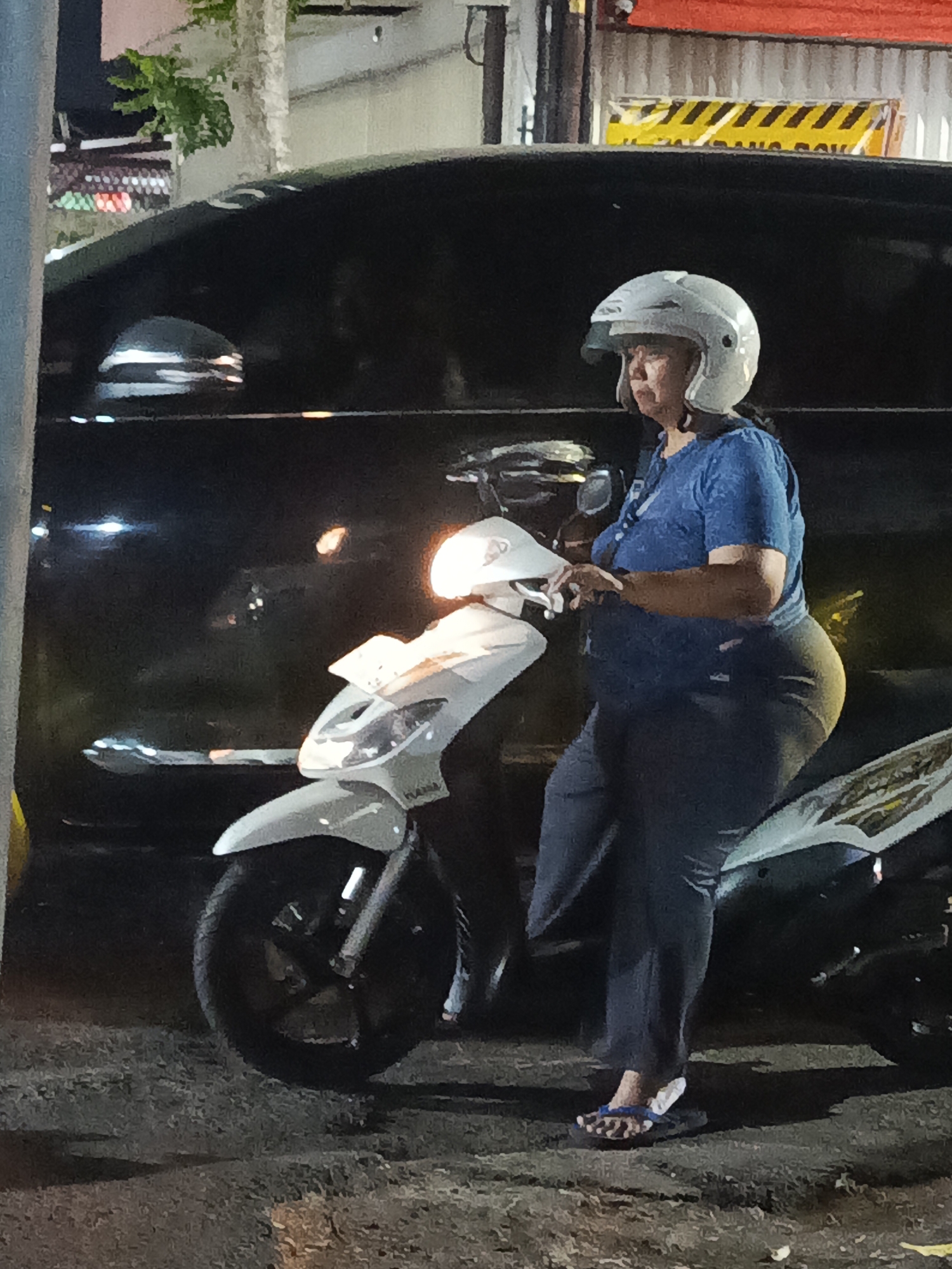 A woman wearing a white helmet and a blue shirt sits on a white scooter. She is on a street in Surabaya, Indonesia, at night. The scooter is parked next to a black vehicle, possibly a car. The background shows a building with a red roof and a yellow and black striped sign. The street is made of asphalt.