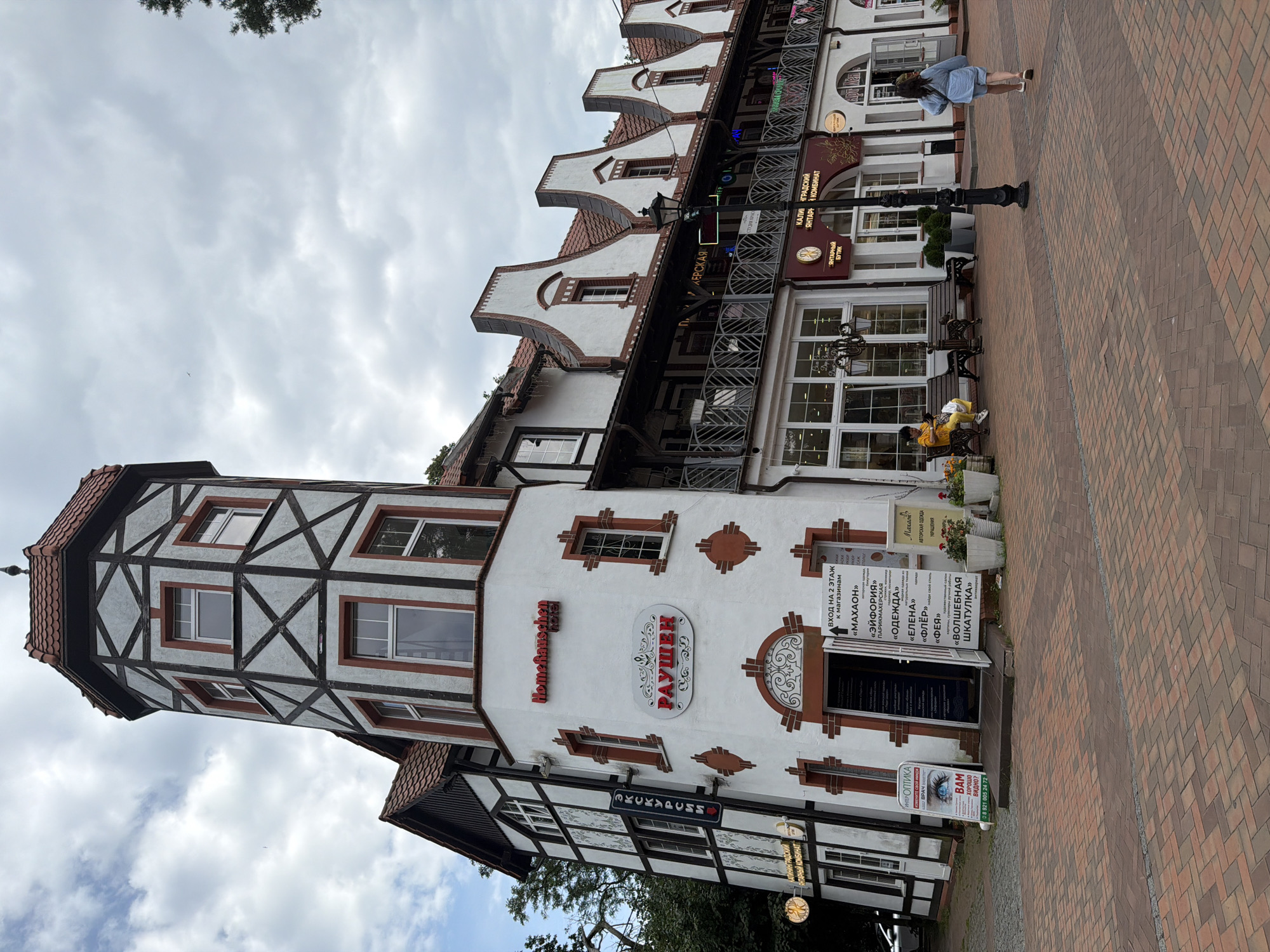 The image shows the exterior of a building in Svetlogorsk, Russia, with a distinct half-timbered tower. The building features a white facade with dark timber framing on the tower and parts of the main building. Red roof tiles are visible on the tower and the gabled roofs of the connected structures.
In front of the building, a brick-paved walkway leads towards the entrance. Several shops are visible with signage in Russian. One sign above the entrance reads "HomeHauschen hotel". Another sign in red letters reads "РАУШЕН".
To the left of the entrance, a standing sign for "ИНТЕР ОПТИКА" displays an image of an eye and text. Near the entrance, there is a small open doorway with a sign listing various services. A woman in a yellow top and white pants sits on a bench outside one of the shops, and a woman in a blue denim jacket and light blue shorts walks away from the camera on the right side of the walkway.
The sky is overcast with clouds, and trees are visible on the left and right edges of the frame. The overall scene depicts a commercial street with architectural elements characteristic of a European style.