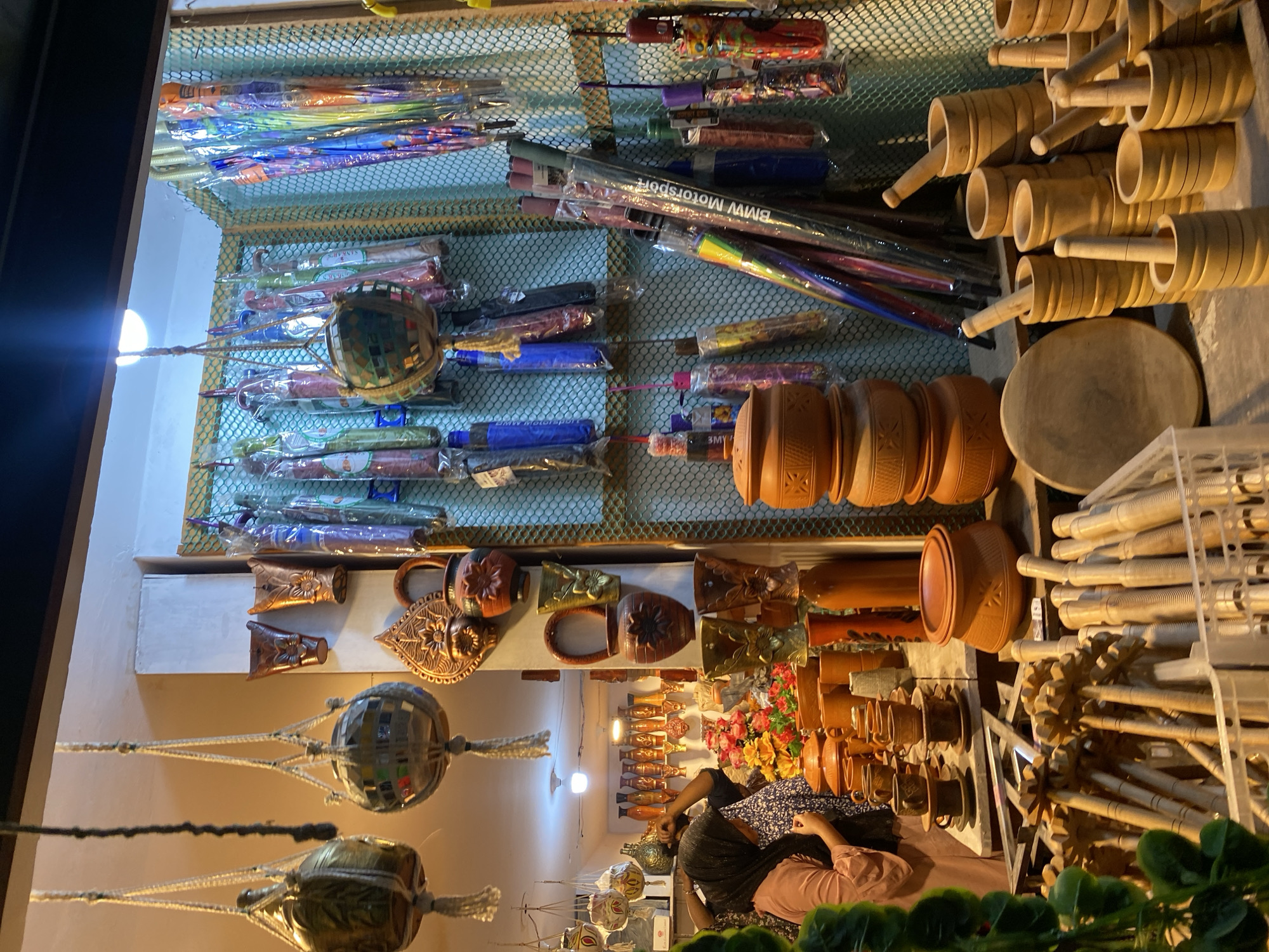 The image captures a stall in Dhaka, Bangladesh, displaying a variety of handcrafted goods. The stall features shelves filled with items, including umbrellas, wooden mortars with pestles, clay pots, and decorative hanging ornaments made of mirrored tiles. A woman wearing a dark scarf and light pink clothing sits behind the stall, seemingly attending to her wares. The stall is brightly lit, highlighting the diverse range of products on display.