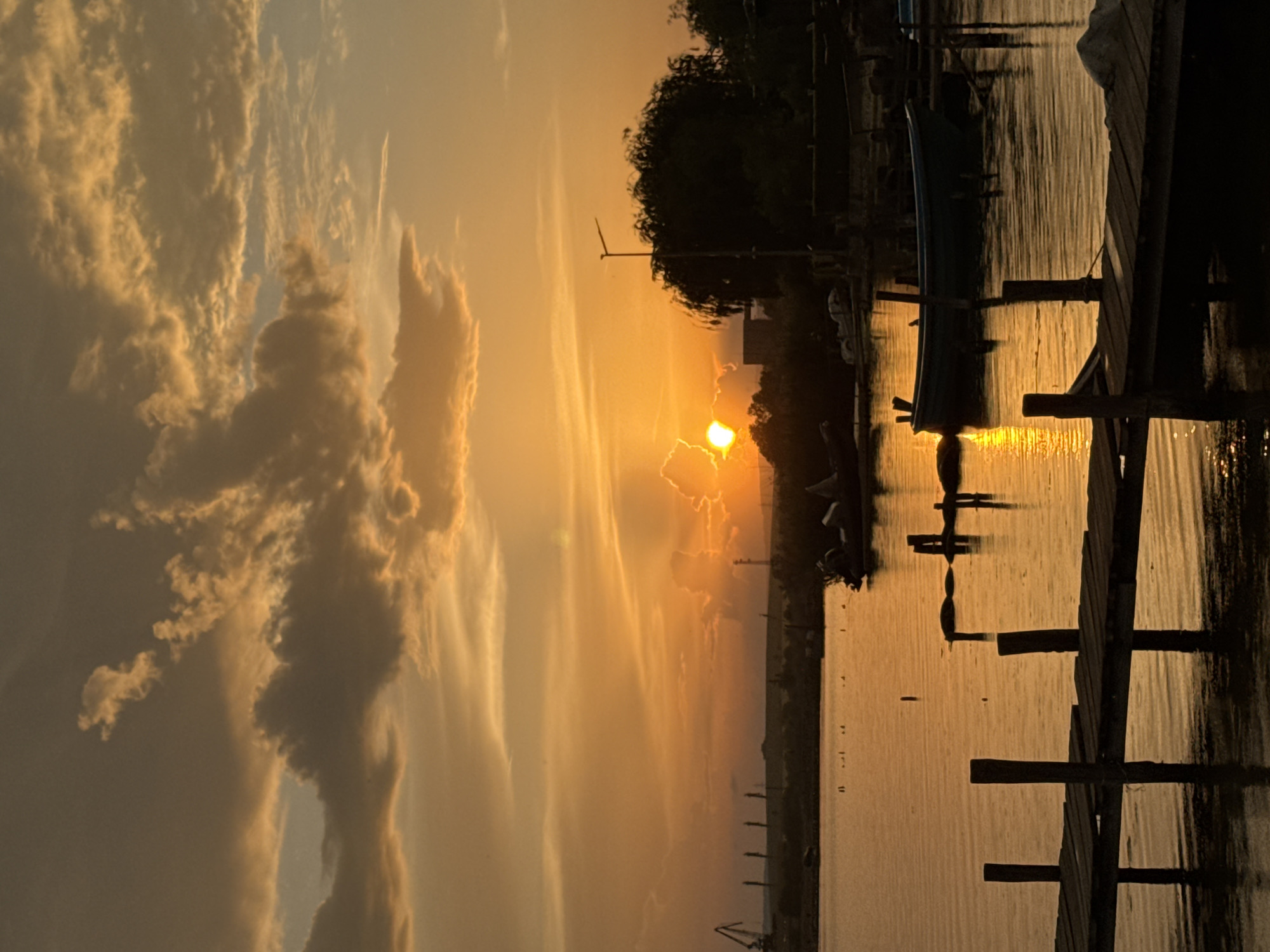 The image depicts a sunset over the water in Казашко, Bulgaria. The sun is setting behind clouds, casting an orange glow on the sky and water. A wooden pier extends into the water on the right. There are boats docked at the pier, and there is a silhouette of trees and buildings in the background.