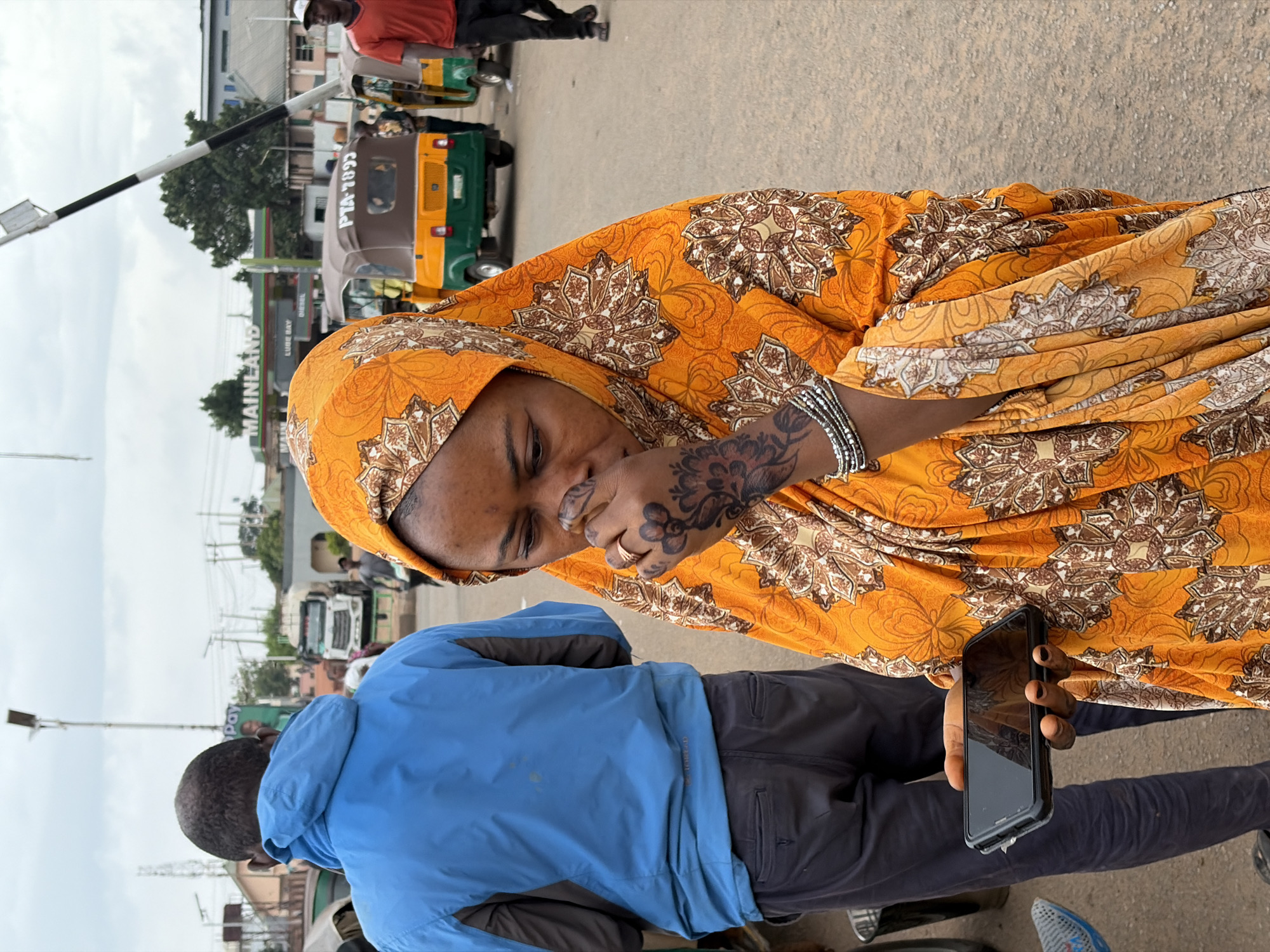 A woman wearing an orange patterned hijab and dress is standing in front of a man wearing a blue jacket and black pants. The woman has tattoos on her left hand and is holding her left hand up to her face. She is also holding a black smartphone in her right hand. The man is turned away from the camera. Behind them, there is a street scene with tuk-tuks (auto rickshaws) and other vehicles. A sign for MAINLAND LUBE BAY is visible in the background. The location is Gura Top, Nigeria.