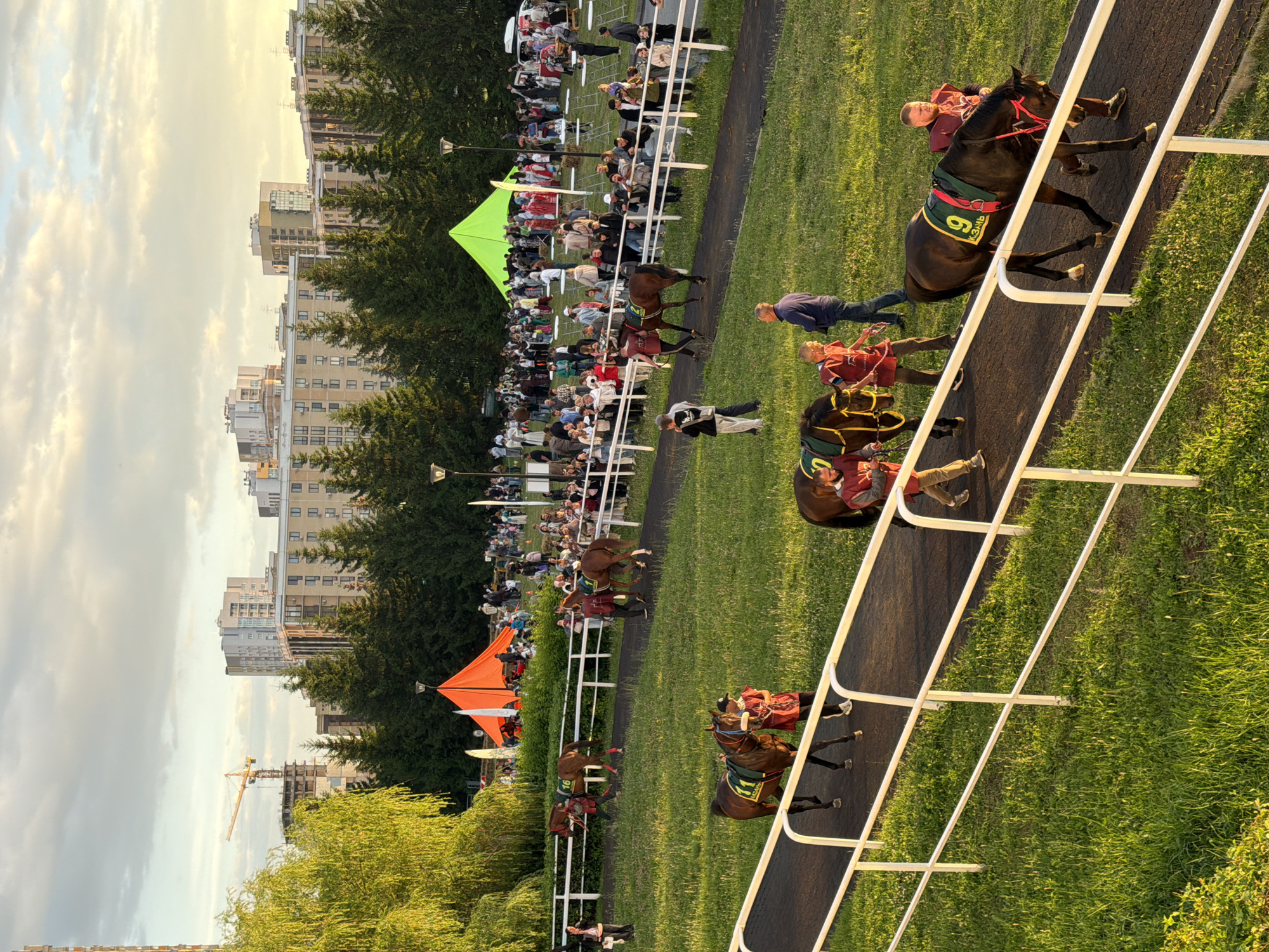 In an outdoor urban setting, a horse race event is underway. The foreground features a racetrack with several horses and their handlers. One horse, with the number 9 on its saddlecloth, is walking along the track, led by a man. Behind it, another horse and handler are visible. The track is bordered by a white fence, and lush green grass lines the sides. In the mid-ground, a crowd of spectators are gathered behind a fence, watching the event. Tents, one orange and one green, are set up in the background, along with trees and apartment buildings. A construction crane is visible in the far distance, suggesting ongoing development in the area. The sky is partly cloudy with a warm glow, indicating either sunrise or sunset. The overall scene depicts a lively event with a blend of urban and natural elements.