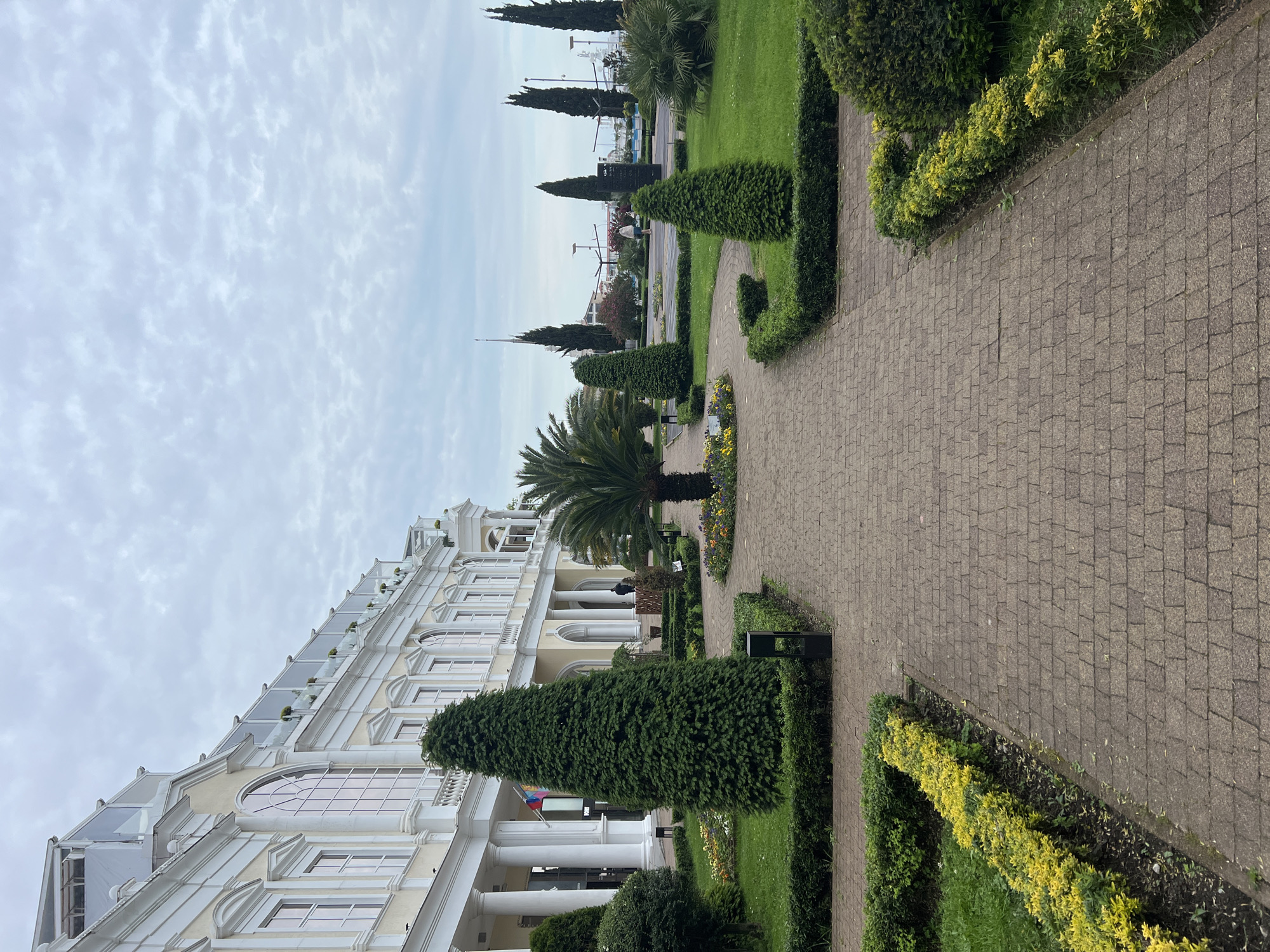 The image depicts a park and building complex in Sochi, Russia, on an overcast day. A brick-paved walkway extends into the distance, bordered by manicured hedges, grass, and flowerbeds. To the left of the walkway, a grand, multi-story building with a classical architectural style features columns and rows of windows. A line of carefully shaped trees, including palms and cone-shaped evergreens, divides the walkway from the building. Distant views show glimpses of the city and waterfront under the cloudy sky.