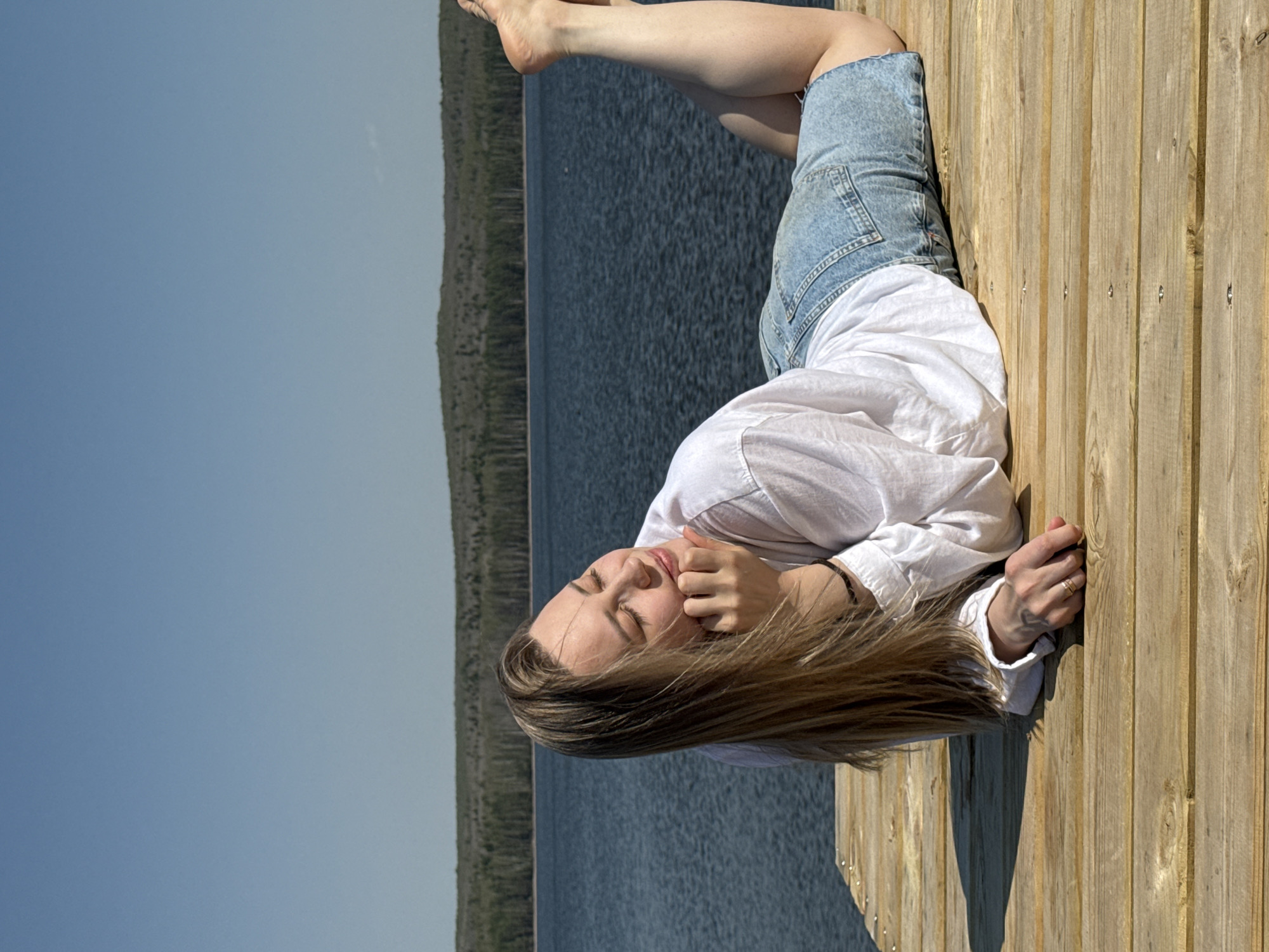 A young woman is lying on a wooden dock, with a body of water and a tree line in the background. The sky is visible in the top left corner of the image. The woman is wearing a white long-sleeved shirt and blue denim shorts. She is resting on her side, with her head resting on her hands. Her legs are bent at the knees, and her feet are slightly raised. The woman has long brown hair, and her eyes are closed. The dock appears to be made of light-colored wood planks, arranged in parallel rows. The body of water is dark blue and has small ripples on the surface. The tree line is green and stretches across the horizon.
The image was taken in the city of Chaikovsky, Russia.