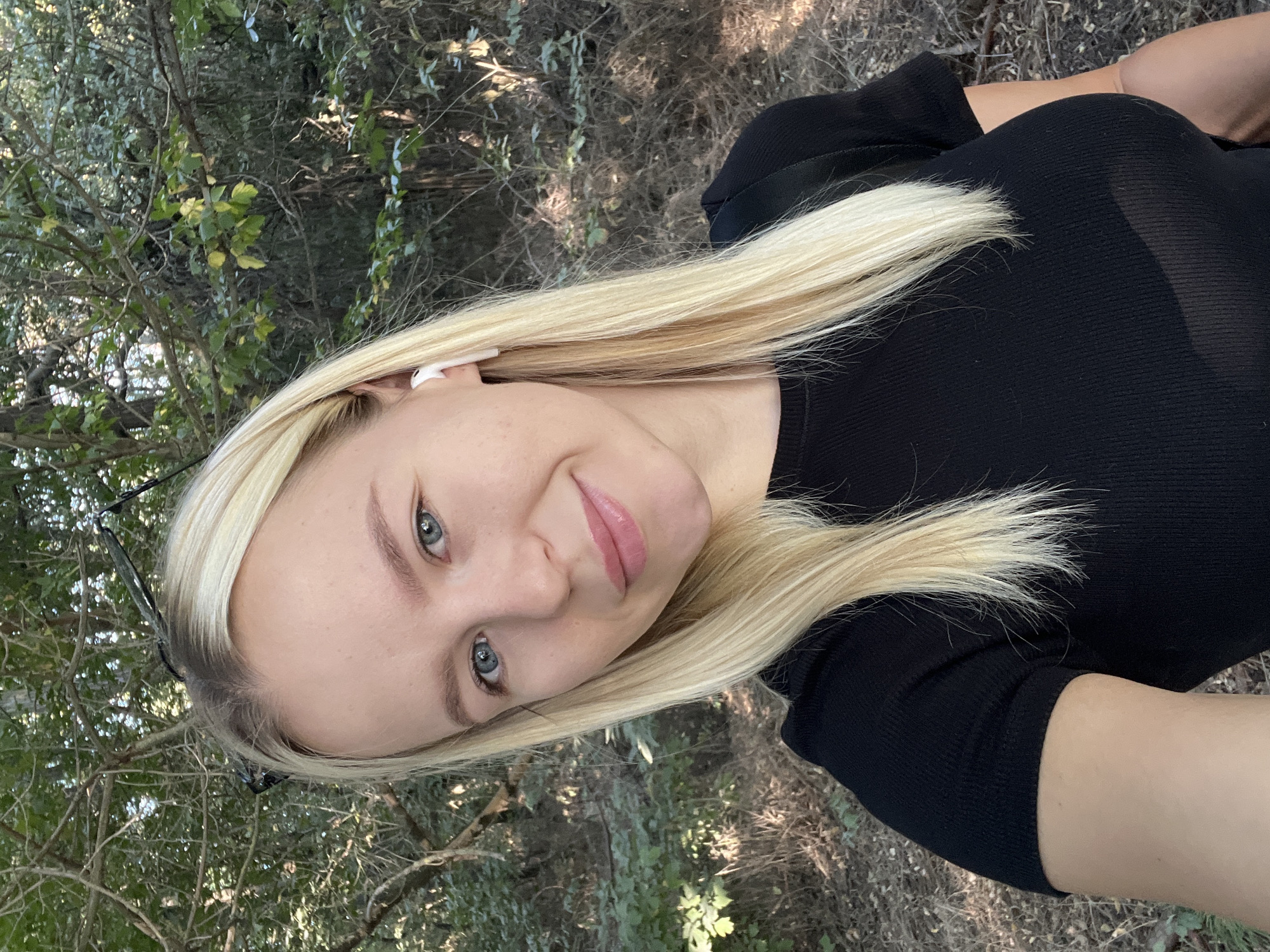 An eye-level, medium outdoor selfie shows a young adult Caucasian woman with long blonde hair and blue eyes. She is smiling and wearing a black t-shirt and white earbuds. The background is a wooded area with trees and foliage.