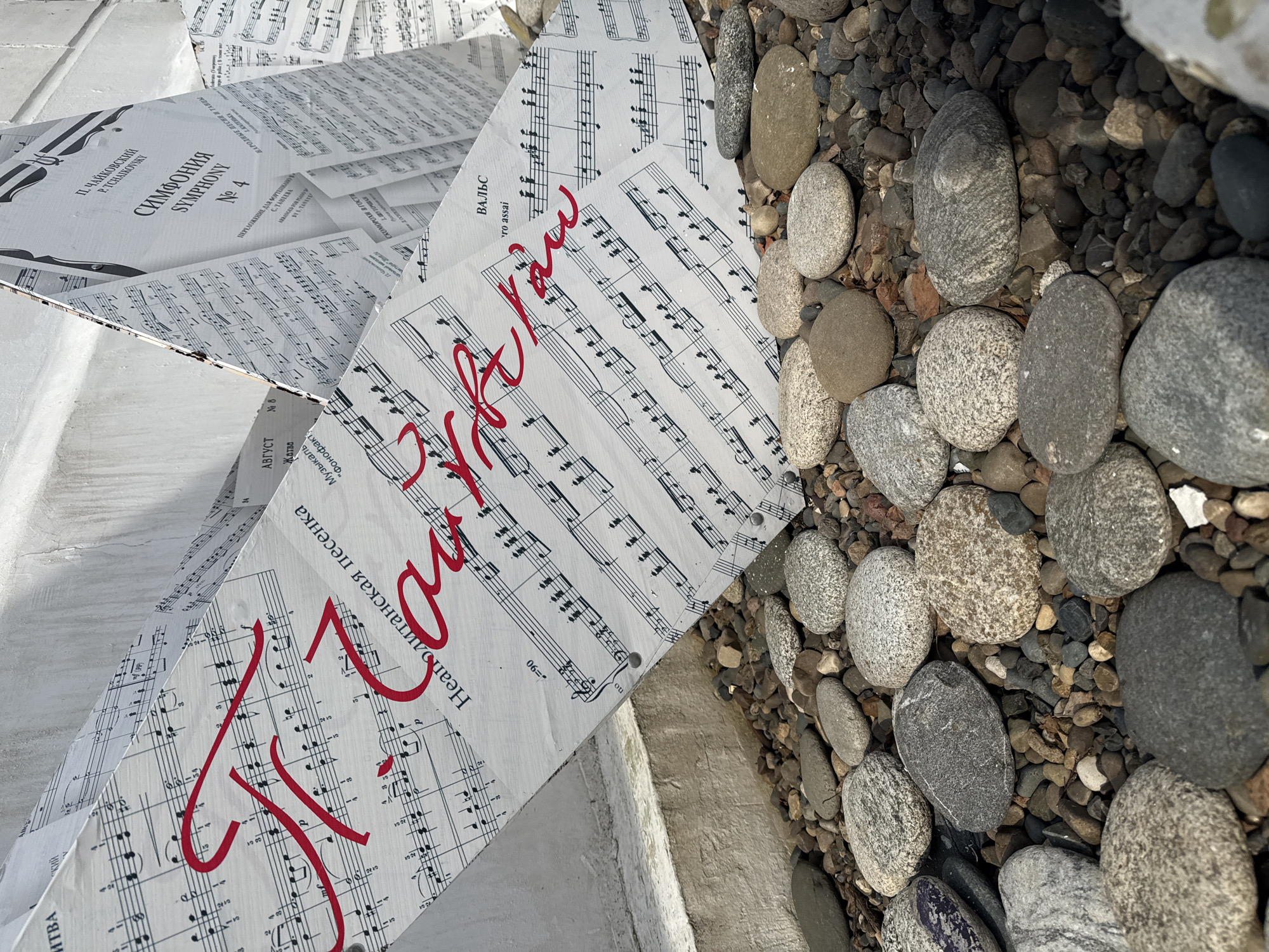 The image captures a low-angle, eye-level view of an outdoor art installation, possibly in a public space, perhaps in Unknown, Unknown. The installation appears to be constructed from multiple white boards or panels printed with musical scores and Cyrillic text. Red cursive script is prominently written across some of the boards, obscuring the notes. The panels form a geometric shape, potentially pyramidal or stepped, and are positioned on a concrete surface next to a bed of smooth, rounded river rocks of varying sizes and shades of gray. The light is soft and diffused, suggesting overcast conditions. There are no visible people in the image. The scene conveys a sense of artistic expression juxtaposed with natural and constructed elements.