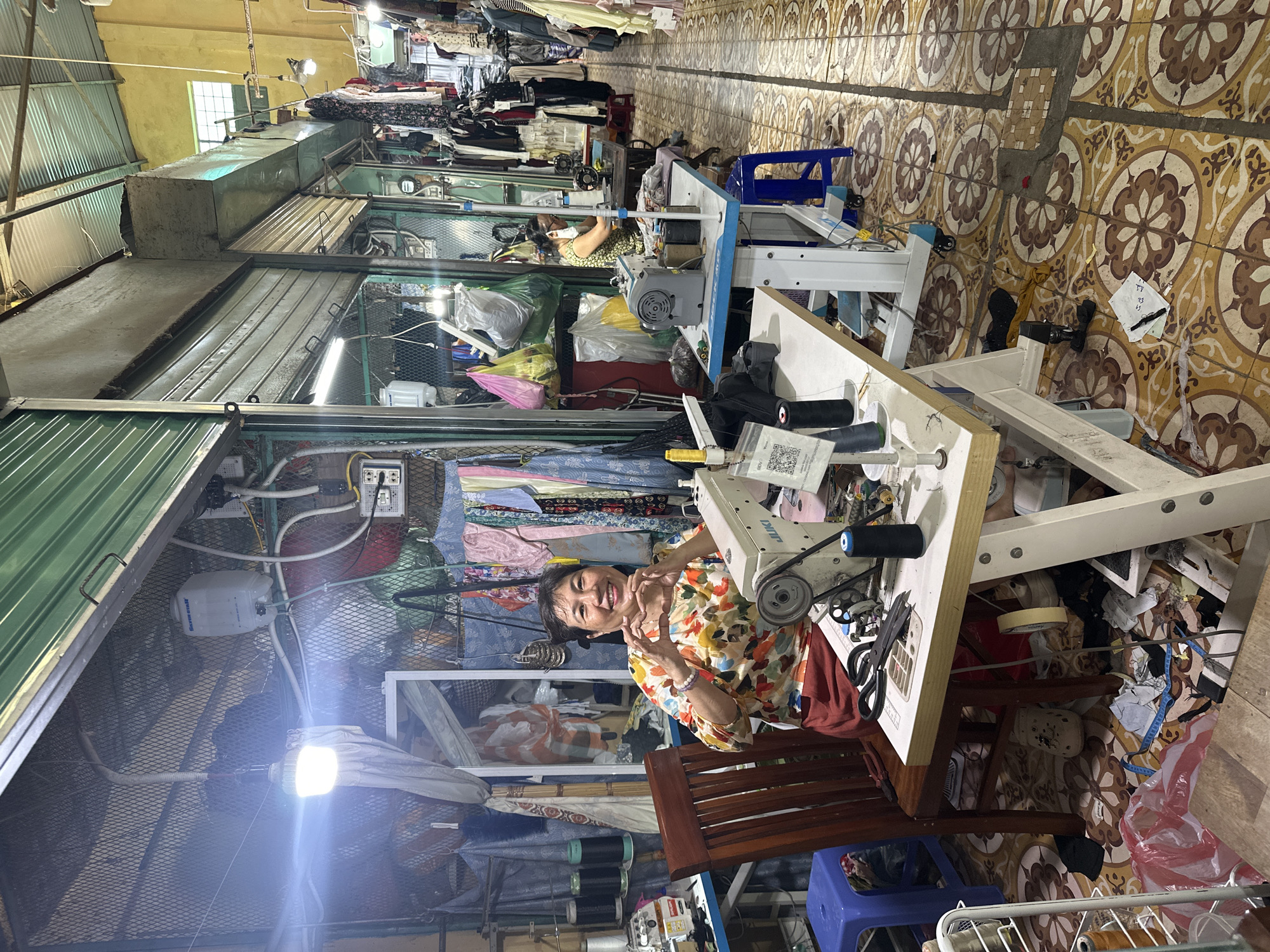 The image depicts a woman seated at a sewing machine inside a small clothing shop. The shop appears to be located in a bustling market area with other clothing stalls visible in the background. The woman is smiling and facing the camera. She is wearing a colorful floral shirt. The sewing machine is on a white table, surrounded by thread, scissors, and scraps of fabric. The stall is filled with textiles, clothes, and sewing supplies. The setting is likely in Đà Nẵng, Vietnam, given the presence of Vietnamese text on some items and the architectural style of the market stalls. The floor is tiled with a patterned design. Artificial lighting illuminates the scene.