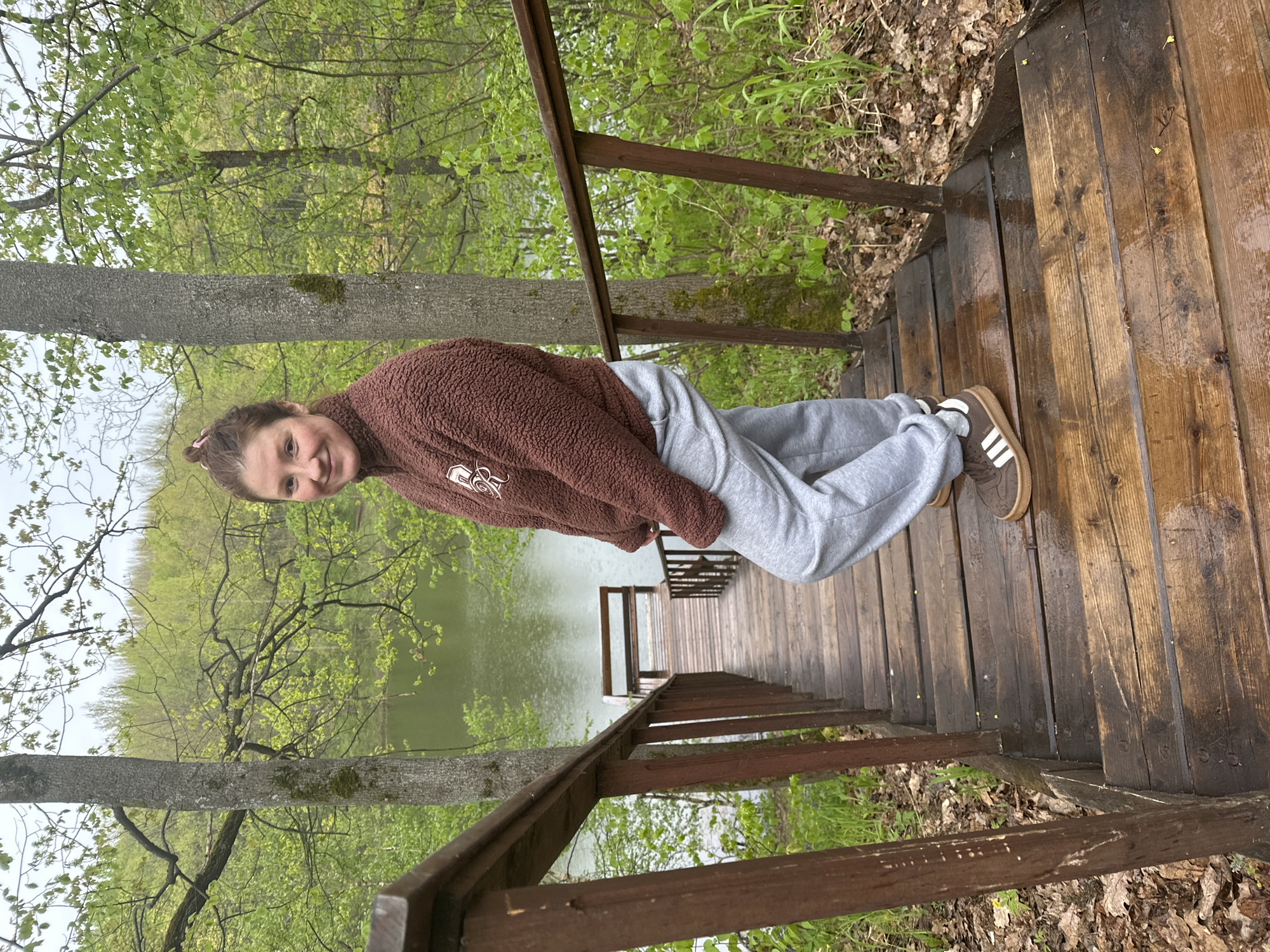 A young woman stands on a wooden staircase descending toward a lake, which is visible through the surrounding trees. She is wearing a brown fleece sweater, gray sweatpants, and brown sneakers with white stripes. The staircase has a wooden railing on both sides, and the steps appear to be wet. The environment suggests a natural setting, possibly a park or wooded area.
The location in the city of Nizhny Novgorod, Russia is not discernable from the image.