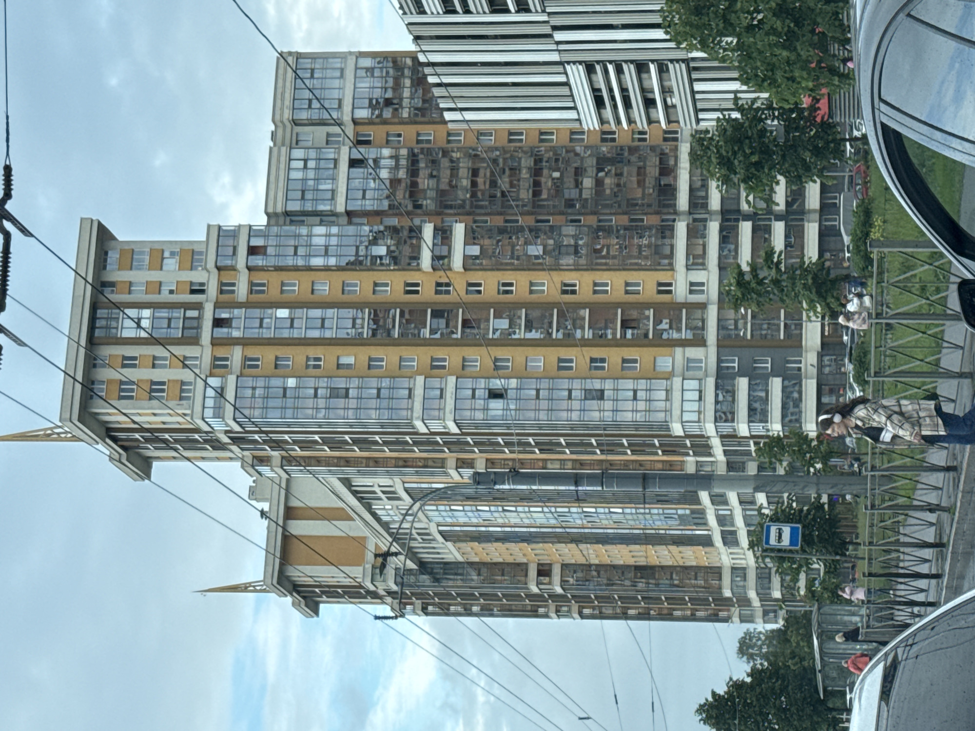 This image captures a view of a tall, modern residential building in Saint Petersburg, Russia. The building features a multi-faceted design with sections of yellow, beige, and gray facades, accentuated by numerous large windows that reflect the cloudy sky. The rooftop is adorned with decorative spires. In the foreground, power lines and a lamppost are visible. To the left of the main building, another structure with a striped facade is present. A bus stop sign is situated near the main building. A woman wearing headphones and a checkered jacket is walking on the sidewalk, while other pedestrians are seen in the background near a bus stop shelter. Green trees line the sidewalk and are interspersed between the buildings. The overall scene suggests an urban environment with contemporary architecture.