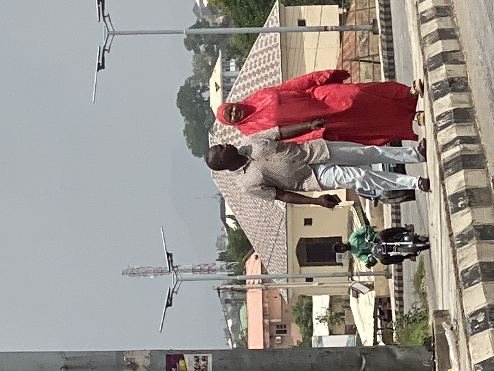 The image captures an outdoor scene in what appears to be an urban or semi-urban environment in Lokoja, Nigeria, viewed from a slightly elevated perspective.
In the foreground, two individuals are prominently featured. A woman, clad in a striking, vibrant red garment that covers her head and body, looks directly towards the camera with a neutral expression. Beside her and slightly in front is a man, seen from behind, wearing a light-colored, patterned short-sleeved shirt and light-toned trousers. He is oriented towards the right side of the frame and appears to be holding a small, dark object, possibly a mobile phone or wallet, in his left hand. The two individuals seem to be walking or standing on a paved surface alongside what looks like a road.
In the mid-ground and background, several low-rise buildings are visible, some featuring distinct patterned roofs. Further along the road to the right, a person wearing a green shirt is seated on a dark-colored motorcycle, which appears to be either stationary or moving slowly. The right edge of the frame reveals a road or pathway defined by a black and white striped curb.
Several tall metal poles with horizontal arms, possibly streetlights or communication antennas, are scattered throughout the scene against a light, hazy, or overcast sky. Patches of green foliage from trees can be seen nestled among the buildings. The lighting indicates it is daytime, though the sky's appearance suggests overcast conditions or atmospheric haze. No discernible text is visible anywhere in the image. The overall impression is a candid snapshot of everyday life in an urban setting.
