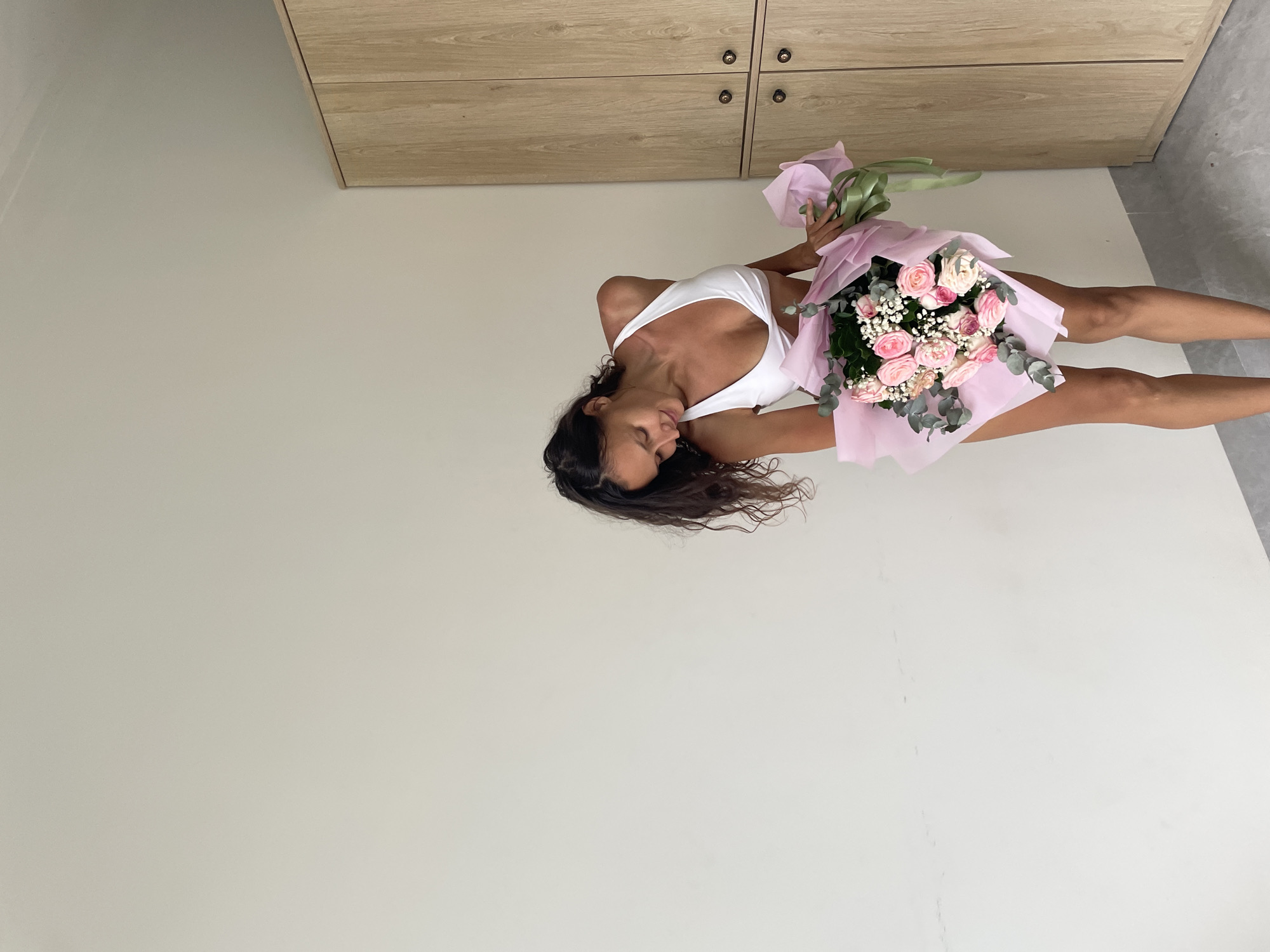 A woman in a white bikini holds a bouquet of pink roses and baby's breath wrapped in pink paper and a green ribbon. She is looking down at the flowers with a slight smile. Behind her is a plain, off-white wall and to her right, a wooden wardrobe with two doors and two round knobs. The floor appears to be made of grey tiles. The scene suggests an indoor setting, possibly a room in a hotel or residence. No specific elements in the image directly indicate a location in Mũi Né, Vietnam.