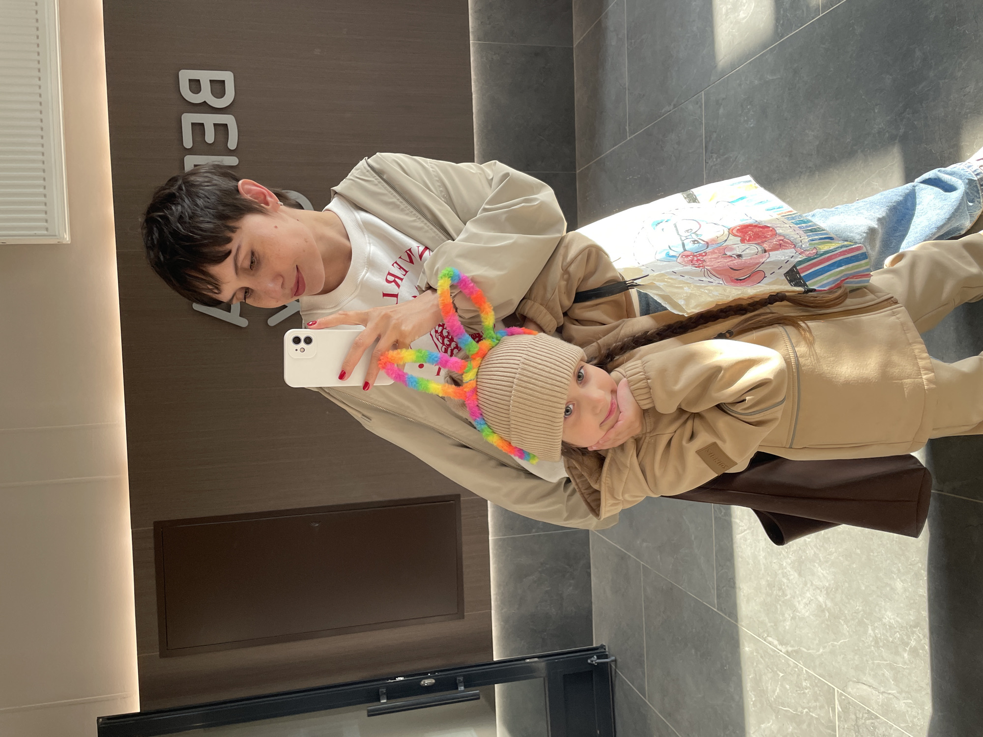 A woman and a young girl are taking a selfie inside a building. The woman, with short, dark hair, is wearing a white shirt with red writing and a beige jacket. The girl is wearing a beige hooded jacket and a beige knit hat with a rainbow-colored headband. The woman is holding a white smartphone. In the background are wood-paneled walls, tiled floors, and an indistinct paper with a drawing. The building's interior includes a door and what seems to be a display case or cupboard. The location appears to be in Izhevsk, Russia.