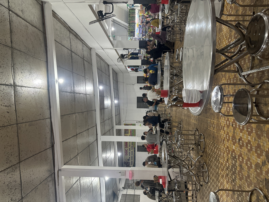 The image depicts an indoor eating establishment located in Nam Hải Lăng Commune, Vietnam. The scene is illuminated by multiple spherical LED lightbulbs mounted on a textured, grey tiled ceiling, which features exposed white beams. The floor is covered in brown and tan patterned tiles. White support pillars are spaced throughout the room, and several black wall-mounted oscillating fans are visible.
Approximately 25-30 individuals, primarily adults, are seated at numerous round and square stainless steel tables, utilizing matching backless stainless steel stools. Many tables feature red plastic caddies containing chopsticks and spoons, along with stacks of white ceramic bowls. Individuals are engaged in dining and social interaction; some are looking at their phones, while others converse.
In the mid-ground, a white pillar displays a partially obscured Vietnamese sign with the prominent text "ĐẶC SẢN" (Specialty), and further text indicating "RƯỢU MINH MANG, RƯỢU BÀU" (Minh Mang Wine, Bàu Wine), suggesting local specialties are offered. In the background on the right, a retail area or market stall is visible, displaying shelves with packaged goods and items hanging from racks. The overall environment suggests a casual, busy eatery or food court setting.