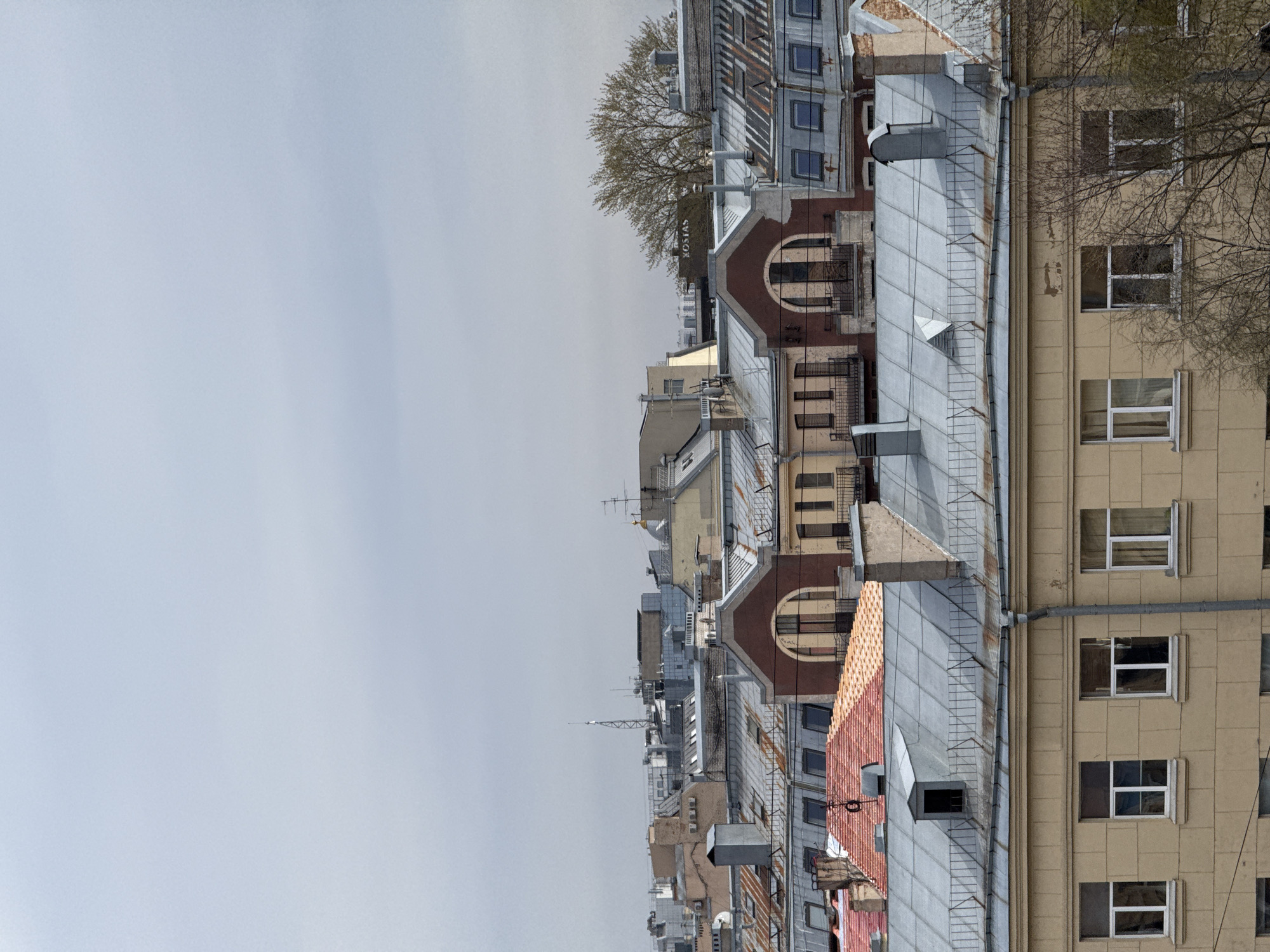 The image shows an aerial view of a dense urban landscape, presumably a city in Unknown, Unknown. The scene is dominated by a variety of rooftops, primarily constructed from grey metal and red tile. The architecture ranges from older, multi-story buildings with arched windows and detailed facades to more modern structures. Many windows are visible, and they vary in size and design. Trees are present, with some showing budding leaves, indicating it might be spring. In the background, there is a pale, overcast sky. The perspective suggests the photo was taken from a high vantage point, such as a taller building.