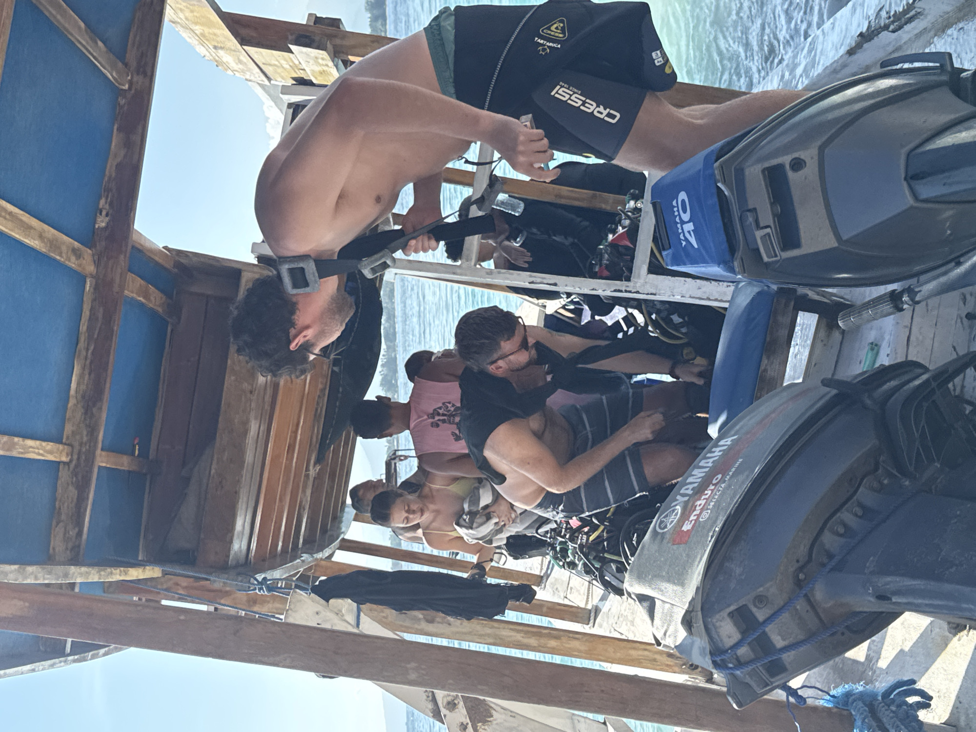 In an outdoor setting in Unknown, Indonesia, a group of people are preparing for a scuba diving activity on a wooden boat. One shirtless man is adjusting his dive mask and gear. Other people are seated, with scuba gear around them. The boat has wooden construction, including wooden support beams and blue panels. The background shows the ocean. Two outboard motors are visible on the boat, one branded "Yamaha Enduro" and another "Yamaha 40".
