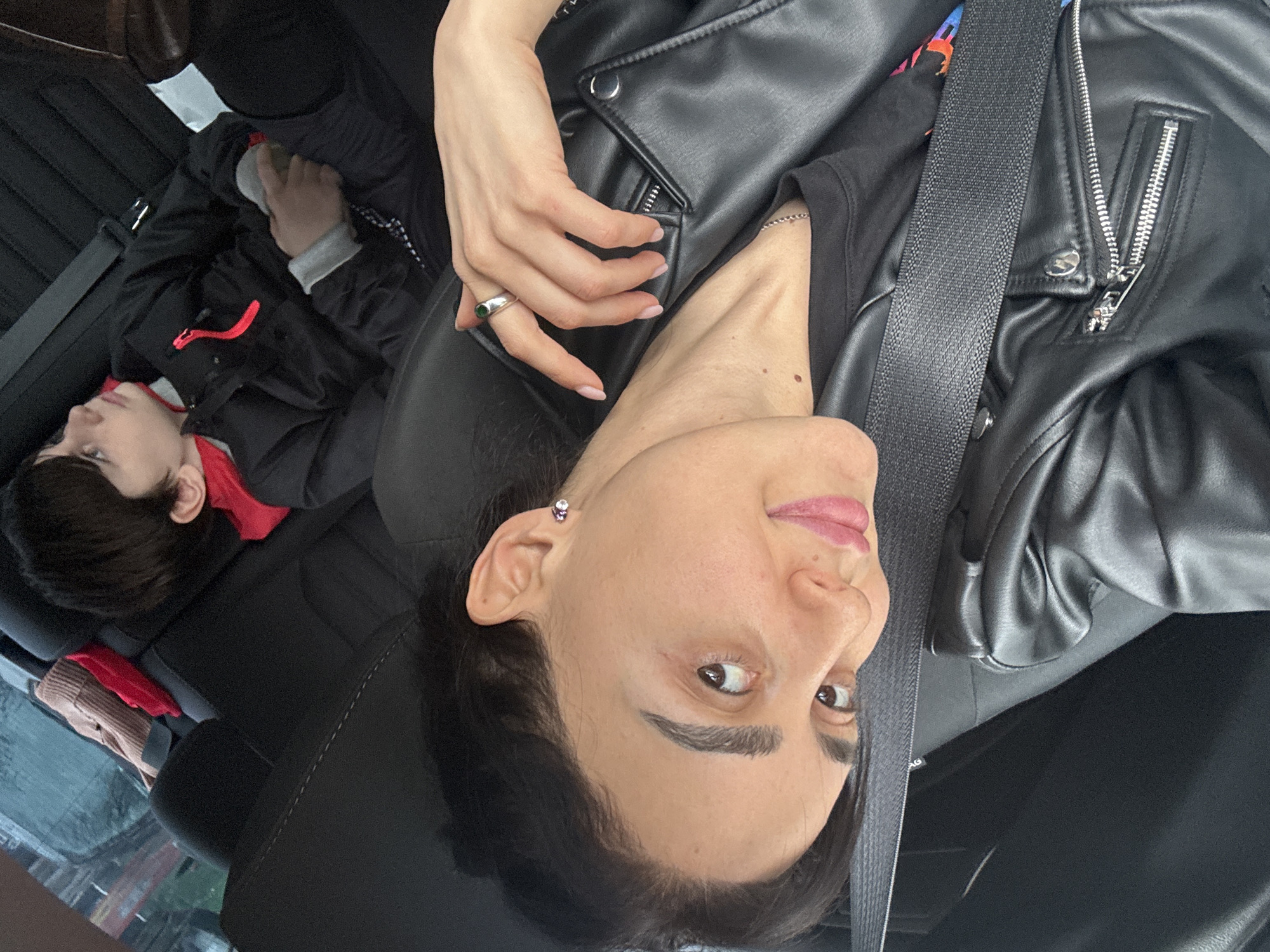 A woman and a child are riding in a car. The woman is in the front seat, wearing a black leather jacket and a seatbelt. She is holding her hand up near her face, showing a silver ring with a green stone. The child is in the back seat, wearing a black and red jacket. The car is in Tbilisi, Georgia. The blurred background through the car window reveals indistinct buildings and streets, indicating an urban environment.