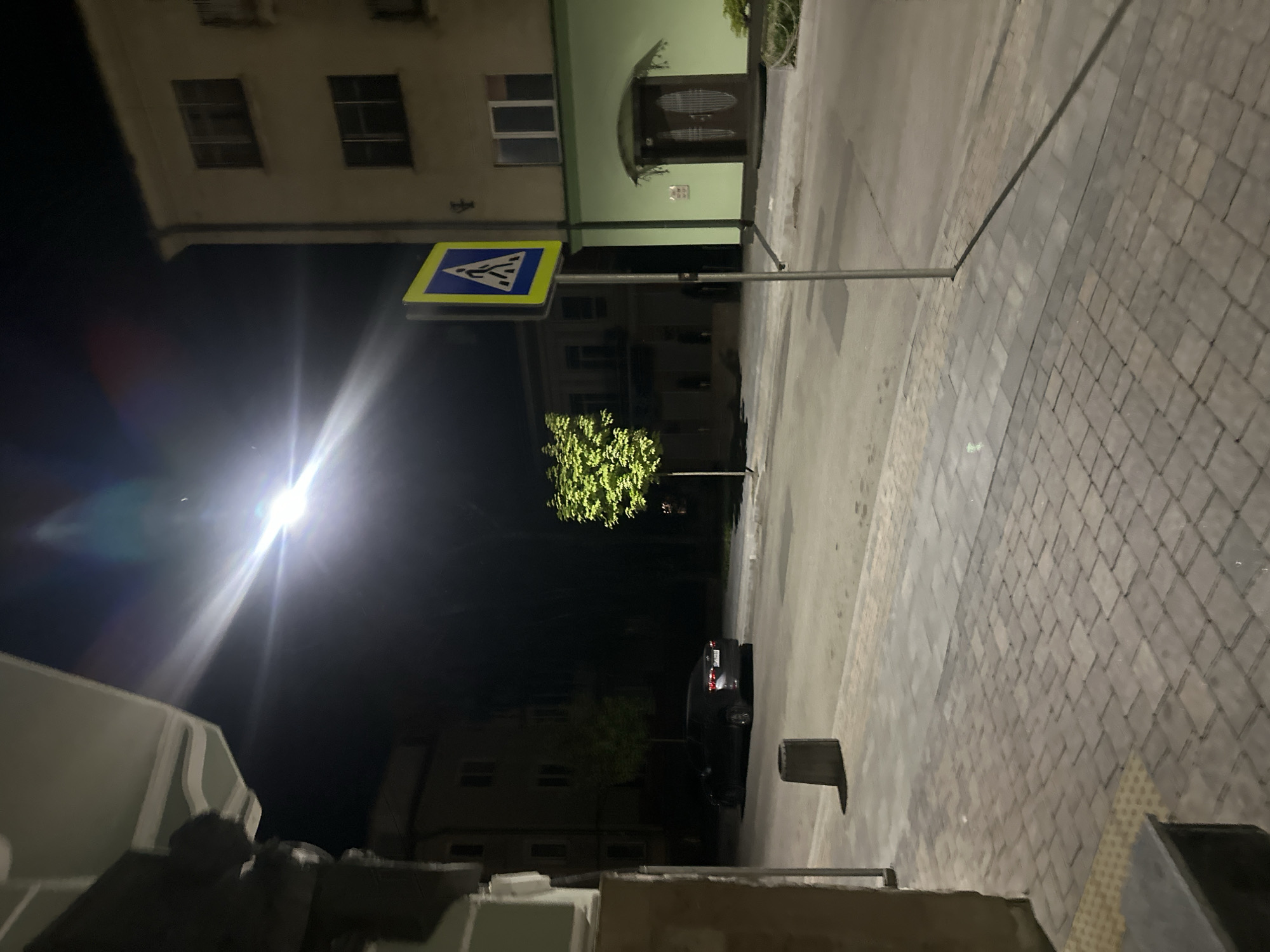 Captured at night, the image depicts a street scene likely in Ternopil, Ukraine, evidenced by the Cyrillic-script pedestrian crossing sign. The scene is oriented vertically with the sky to the left and ground to the right. A brightly lit pedestrian crossing sign stands on the left side of the street, illuminating a tree behind it. The street itself is made of light-colored pavement stones. A car is parked further down the road beyond the tree. Buildings line both sides of the street, some with lit windows, suggesting residential or commercial occupancy. The dominant feature is the pedestrian crossing sign with its reflective surface.