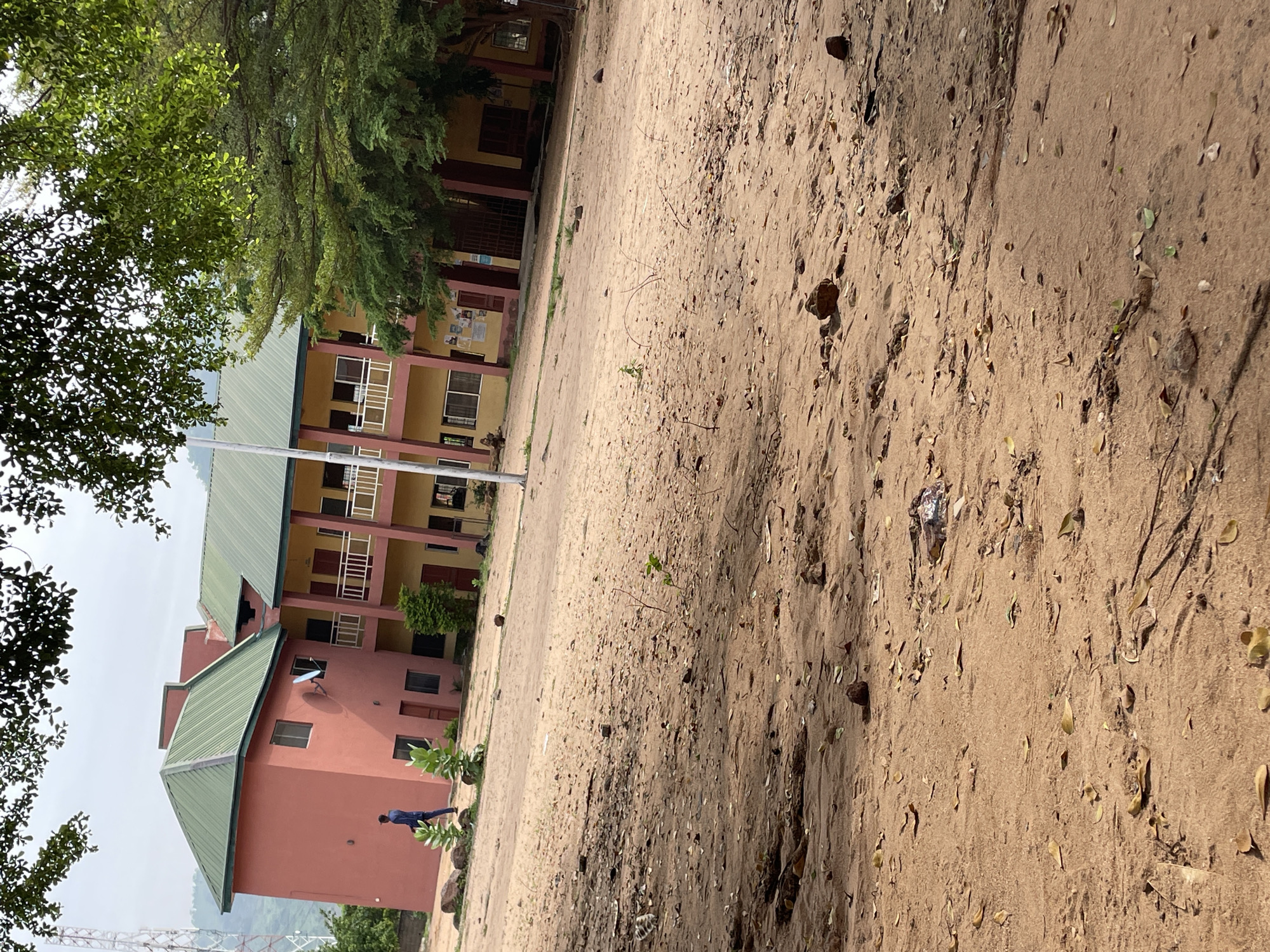 In Lokoja, Nigeria, an outdoor scene depicts a two-story building with a red and yellow facade and a green roof. The building appears to be a school or administrative complex, with multiple windows and balconies. In front of the building, a person in a dark blue outfit is walking across a sandy area littered with leaves and small stones. Trees with lush green foliage overhang the scene, casting shadows on the ground. In the background, a radio mast is visible on a distant hill, and the sky is overcast. The overall impression is one of an academic or governmental institution in a somewhat arid, natural environment.
