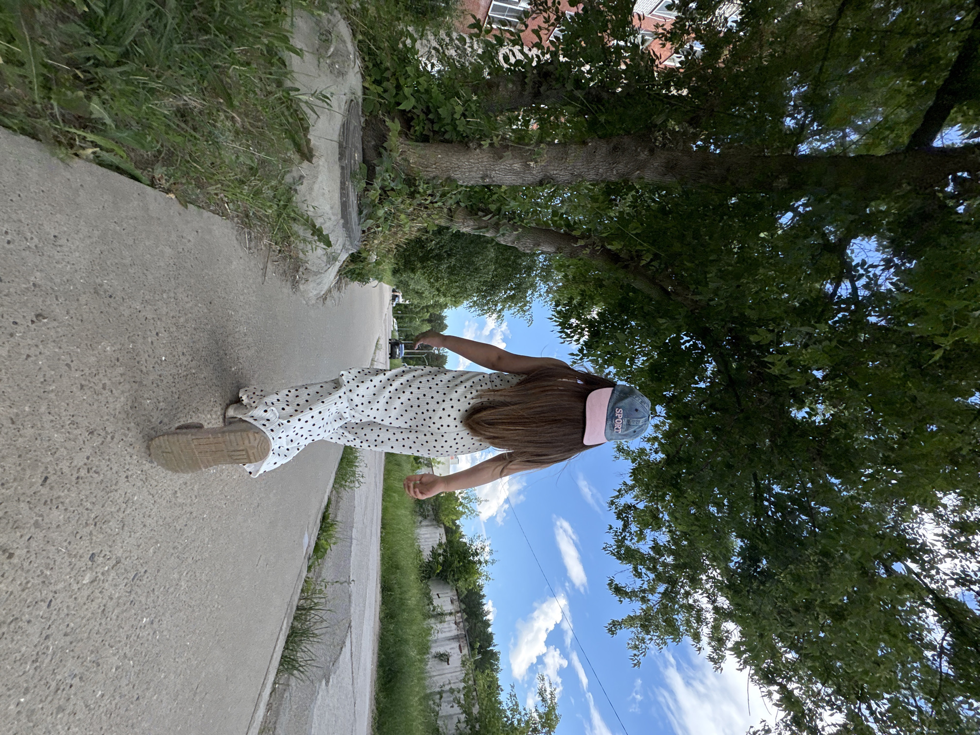 The image shows a young girl from behind, walking on a paved sidewalk. She is wearing a white dress with black polka dots and a light blue baseball cap with pink trim and the word "SPORT" embroidered in white. The girl's long, brown hair is visible. Her right arm is extended outwards. She is wearing light-colored slip-on shoes.
The setting appears to be an urban or suburban area, with a sidewalk bordering a grassy area and trees. A large tree with a thick trunk is prominent in the foreground, its branches and leaves extending over the sidewalk and partially obscuring the sky. In the background, there is a building with multiple windows, and a cloudy blue sky. The image conveys a sense of a casual, outdoor activity, likely during the daytime given the bright sky. The lighting suggests it is daytime, and the presence of green foliage indicates a warmer season.
The scene is set in Chaikovsky, Russia.