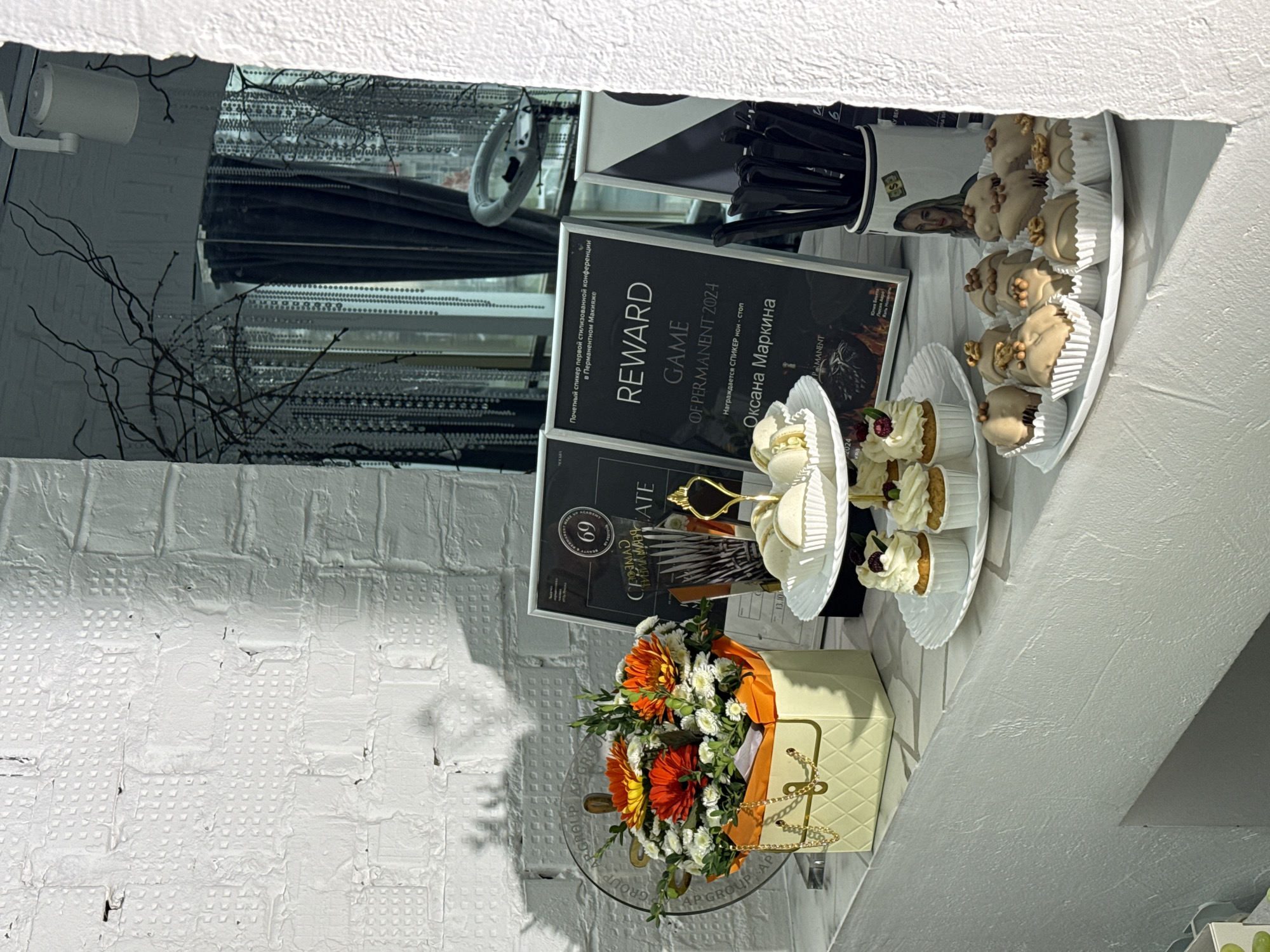The image shows a shelf display in what appears to be a business in Chaikovsky, Russia. The display includes several objects. There is a white brick wall in the background. A flower arrangement, consisting of white and orange flowers, is displayed on a glass plate. A small, handbag-shaped box is also present. Several desserts are placed on white platters, including small cakes and other pastry items. A black certificate framed in white has Russian text. A second framed certificate is placed behind the first, the text reads "Reward Game Of Permanent 2024." Other objects, including a pair of sunglasses and a container full of black pens or markers are displayed near the certificates and desserts. A security camera can be seen.