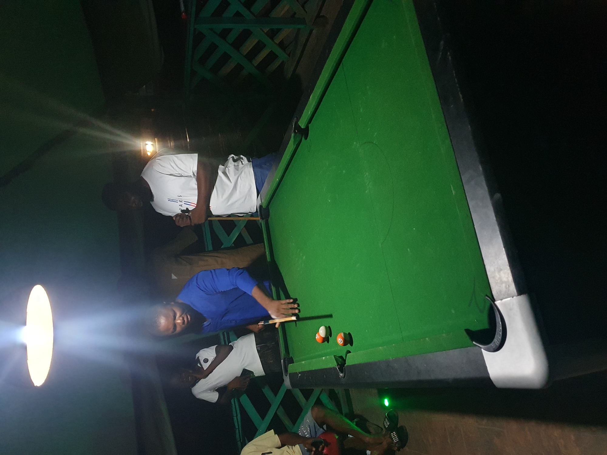 At night, a group of men are playing billiards on a green felt table located outdoors, likely on a patio or rooftop in Lokoja, Nigeria. A bright overhead light illuminates the scene. One man in a blue shirt is actively playing, poised to strike a ball with a cue. Two other men, one in a white t-shirt with "PARIS" printed on it and another in a white shirt with a black collar, stand nearby observing or waiting their turn. Another individual in a yellow shirt is partially visible on the left, wearing flip-flops. The environment appears to be a leisure or entertainment area with a wooden railing in the background.