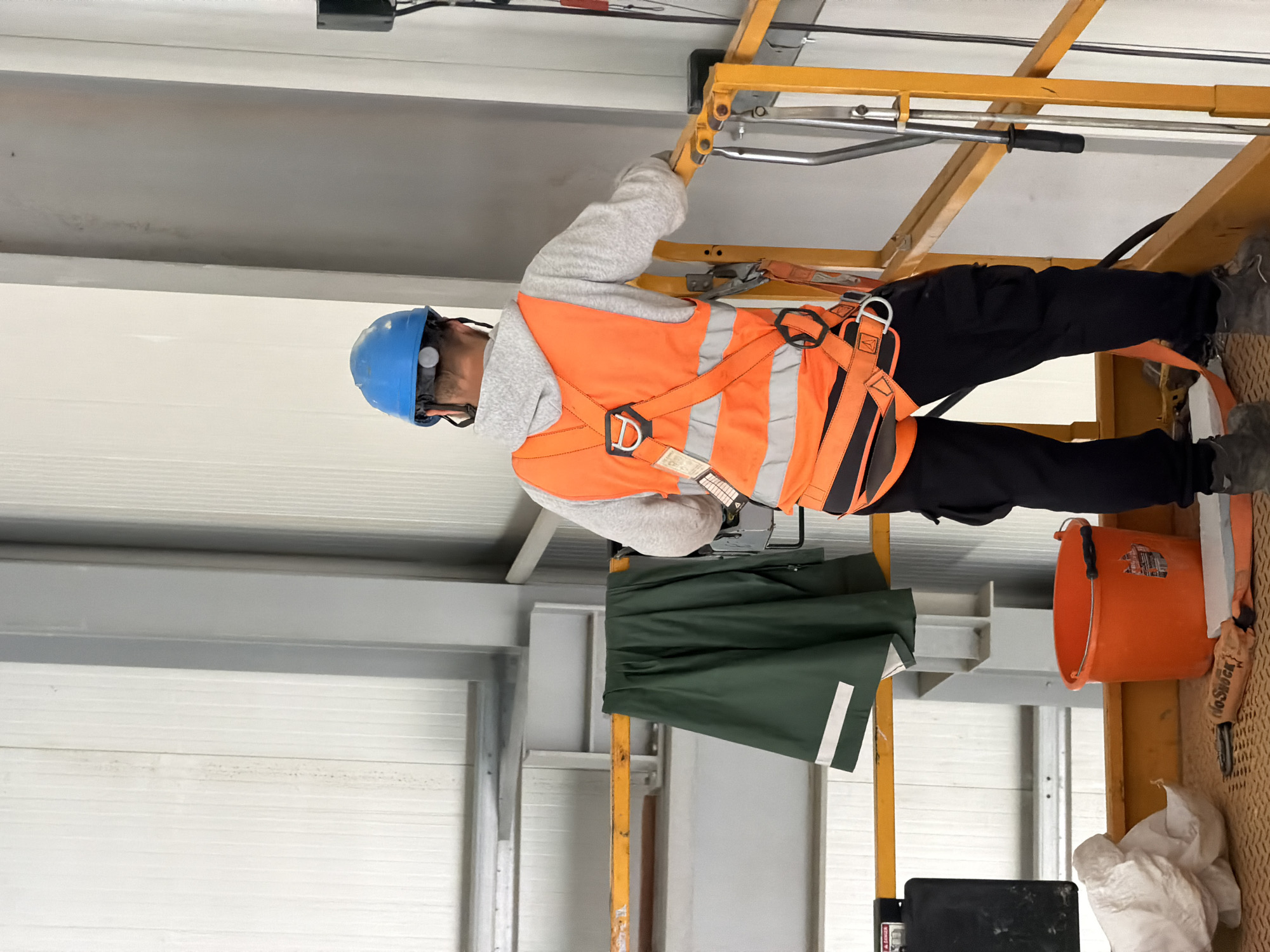 A male construction worker, wearing a blue hard hat, a grey hoodie, and an orange safety vest with reflective strips, stands on a yellow metal platform. The worker is looking away from the camera, towards a white corrugated wall. A dark green rain jacket hangs on a yellow railing to the left of the worker. In the foreground, an orange bucket, a white bag, and some construction materials are visible on the platform. The environment appears to be an indoor industrial setting, possibly a construction site. The scene is devoid of any specific Janowice landmarks.
