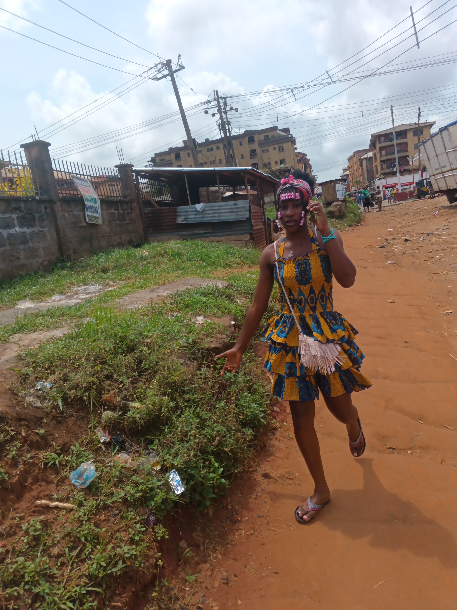 A young woman is captured in a medium shot, walking on a dirt road in Onitsha, Nigeria. She is wearing a vibrant yellow and blue patterned dress with ruffles and a white fringe bag slung across her body. A pink headband with pink decorative elements adorns her head. She is holding a mobile phone to her ear, engaged in a conversation. Her left arm is extended outwards. The road is unpaved and reddish-brown, with scattered debris along the sides. To the left of the road, there is a patch of overgrown green grass and more discarded trash. In the background, several buildings, including a multi-story structure, are visible. Power lines and utility poles run across the sky. A white truck is parked further down the road. The sky is overcast with visible clouds.