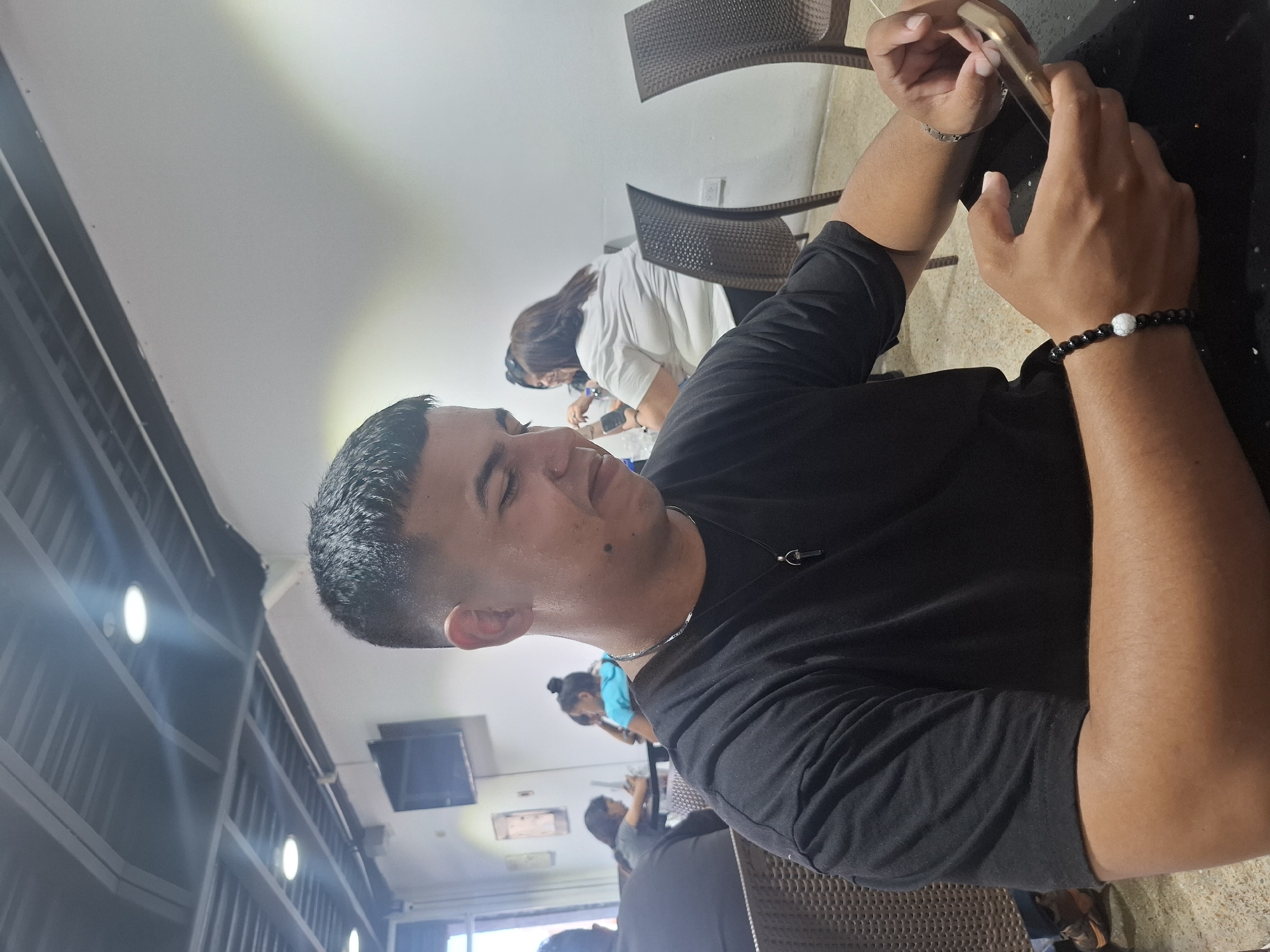 A close-up, slightly rotated photograph depicts a man in a black t-shirt holding a phone. He has short, dark hair and wears a bracelet on his right wrist. A woman in a white shirt and another person in a blue shirt are visible in the background. Other people are seated and visible. The setting appears to be an indoor seating area in a restaurant or cafe, possibly in Juan Griego, Venezuela, given the prompt's instruction. The background also includes a window and some shelving with lights.