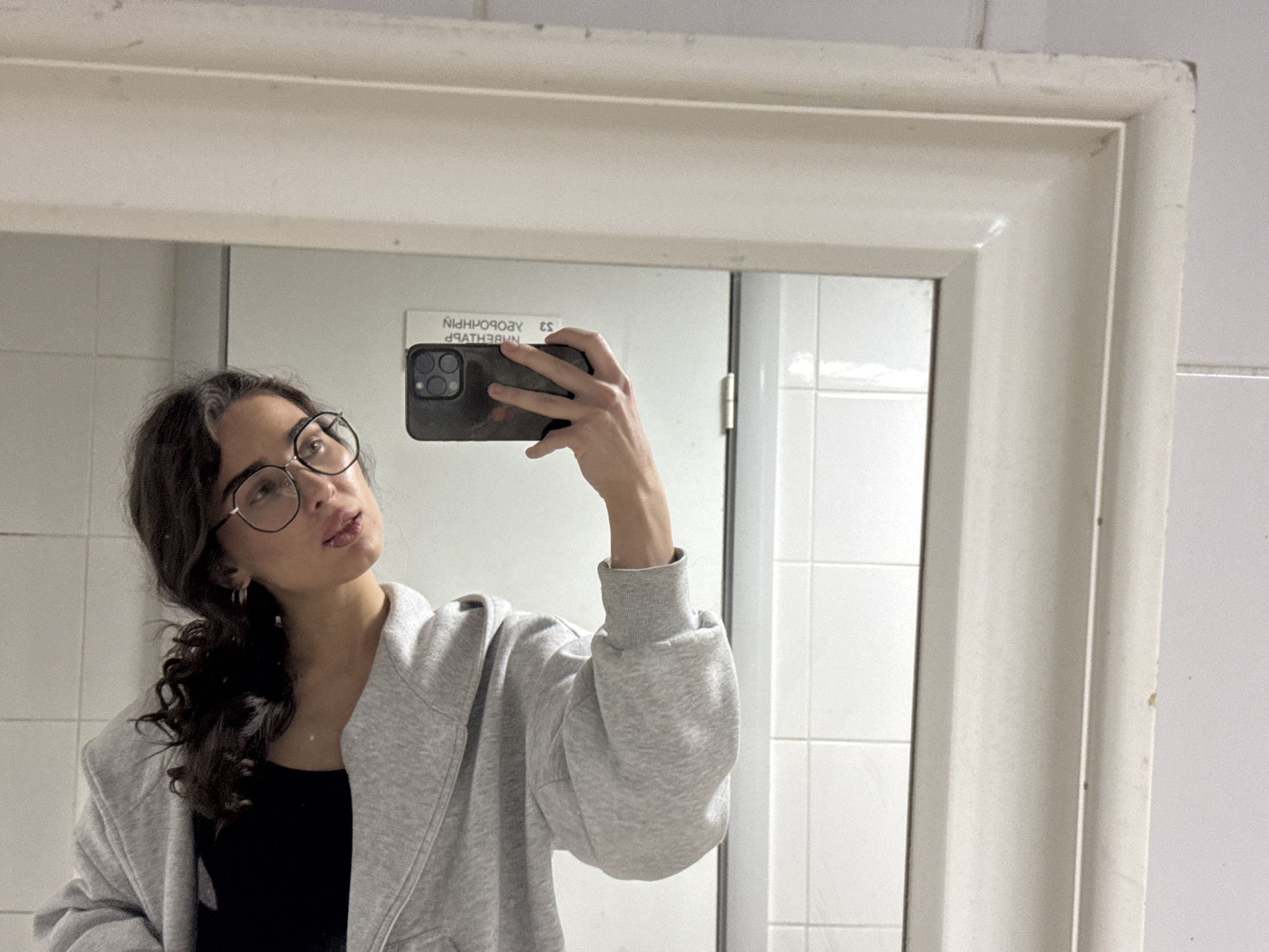 A young woman with dark, curly hair pulled back and wearing round glasses, a black top, and a grey zip-up hoodie is taking a selfie in a mirror. Her head is tilted back, and her lips are slightly parted. The mirror is framed by a beige border, and the background appears to be a tiled wall. The phone in her hand is black, and its screen is visible. A sign with text in Cyrillic script is reflected in the mirror behind her. The scene is likely a bathroom in Kazan, Russia, given the text on the sign.