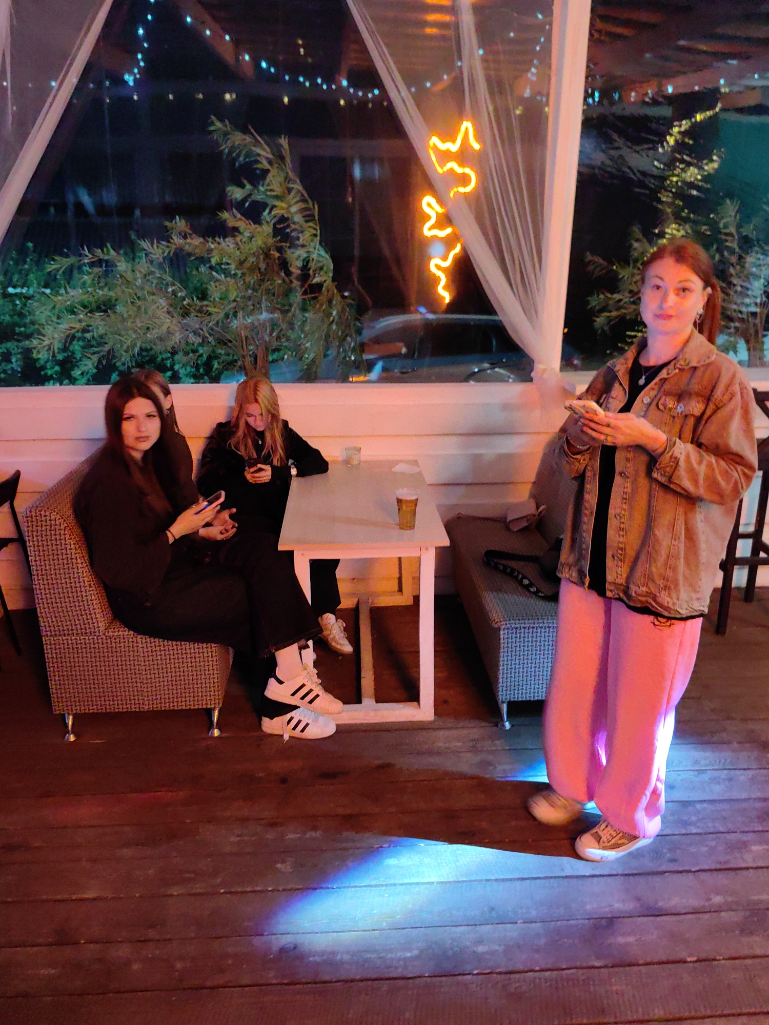 The image depicts an outdoor seating area, possibly a cafe or restaurant, with a wooden floor. Three young women are present. Two of them are seated on a patterned sofa, each engrossed in their mobile phones. The woman on the left is wearing a dark outfit, and the woman on the right has blonde hair and is wearing a black top. A white table is positioned in front of them with a glass of amber liquid on it. To the right, another young woman stands, holding a phone in her hand and wearing a denim jacket over a black top and bright pink trousers. Behind the women, a window or opening reveals a nighttime scene with illuminated string lights and some greenery. Neon light, shaped like a squiggle, is visible on the right side of the frame, behind a sheer curtain. The overall lighting suggests an evening setting, with warm tones and some colored light reflections on the floor. The location appears to be in a natural setting given the greenery outside, and it is stated to be in Нарацкі сельскі Савет, Belarus.