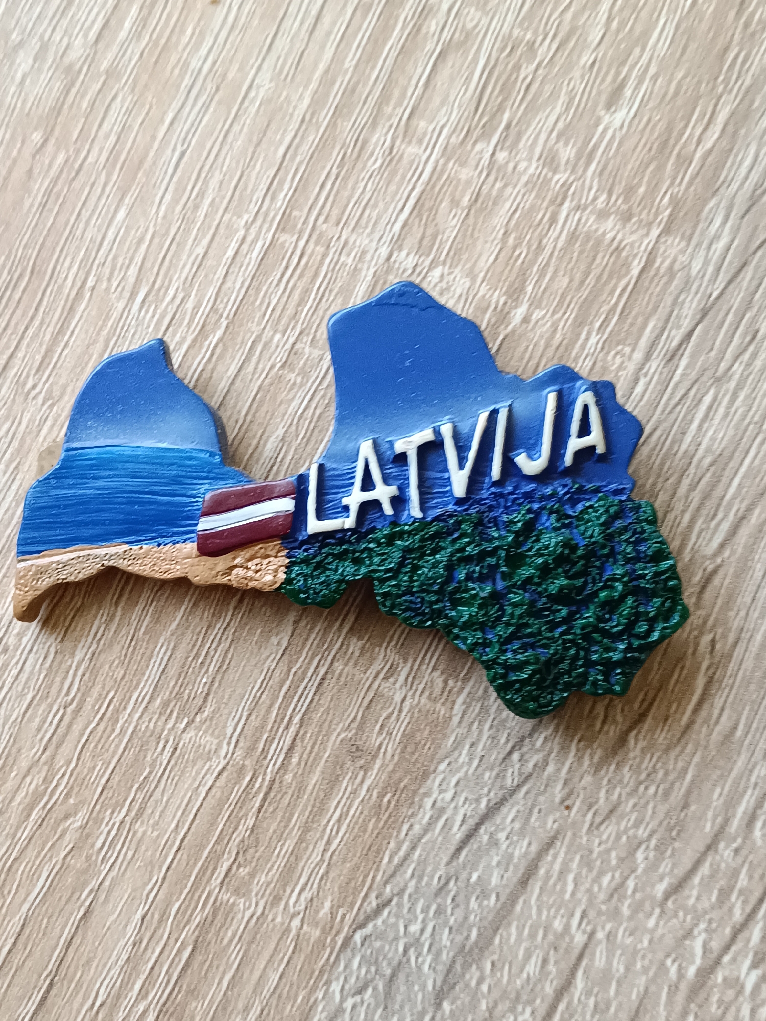 The image displays a magnet shaped like the outline of Latvia, placed on a light-colored wooden surface with visible grain. The magnet features a blue background representing the sky and water, with sandy textured areas depicting a beach. The Latvian flag is incorporated into the design, positioned on the lower left side. White lettering spells out "LATVIJA" across the upper portion of the magnet, overlaid on a blue section that transitions to a textured green area, likely representing forests. The object is photographed from a slightly elevated, angled perspective. There is no indication of the city of Tebras, Latvia, or any people in the image.