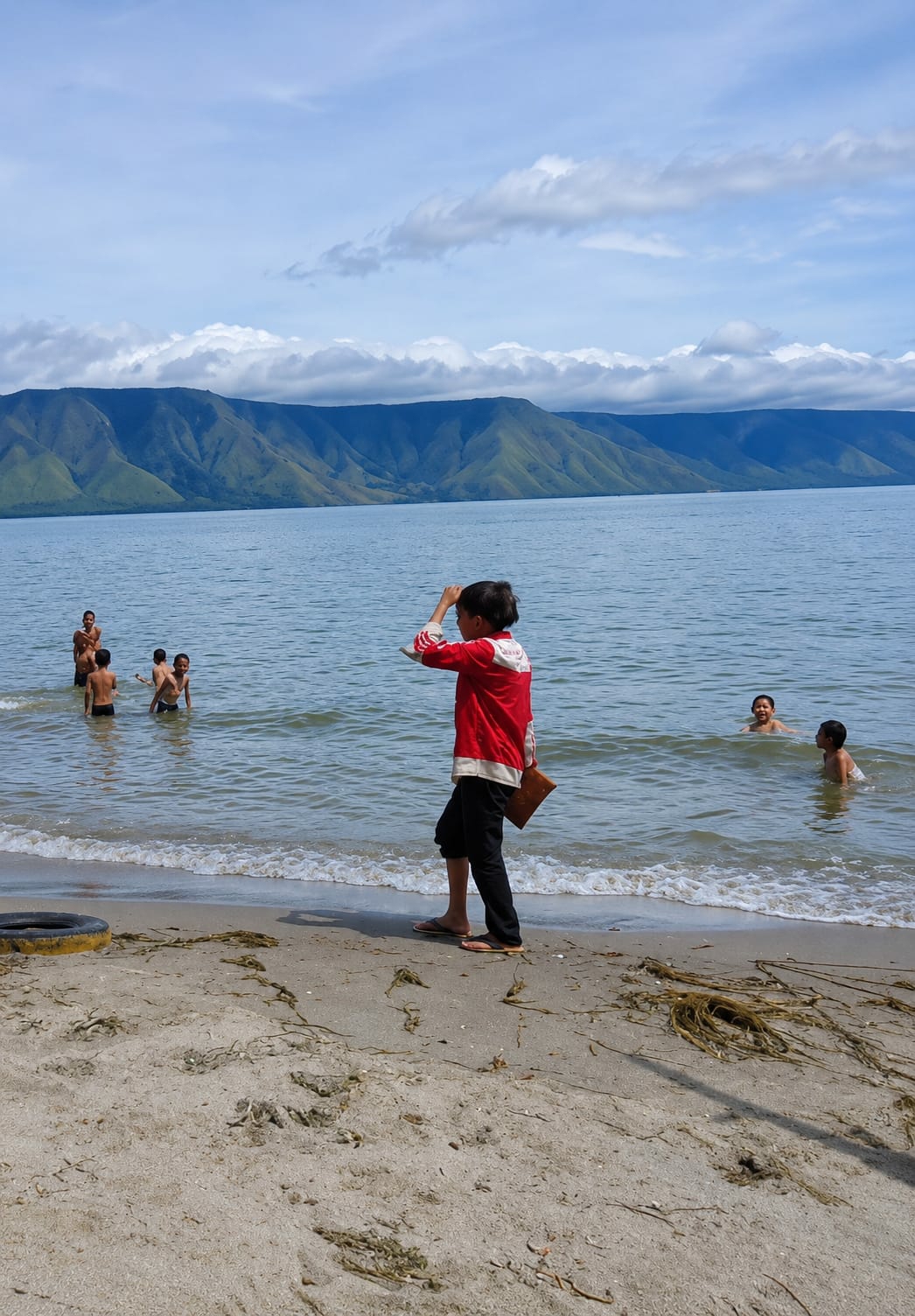 In Situngkir, Indonesia, on a bright, cloudy day, several young people are enjoying the water of a large lake. In the foreground, a person wearing a red jacket with white trim and black pants stands on a sandy beach, shielding their eyes from the sun with one hand and holding a brown object. Waves gently lap at the shore, where dried vegetation and a discarded tire are visible. In the background, a group of boys are playing in the shallow water. Beyond the lake, lush green mountains rise, forming a majestic backdrop under a sky filled with dramatic clouds.