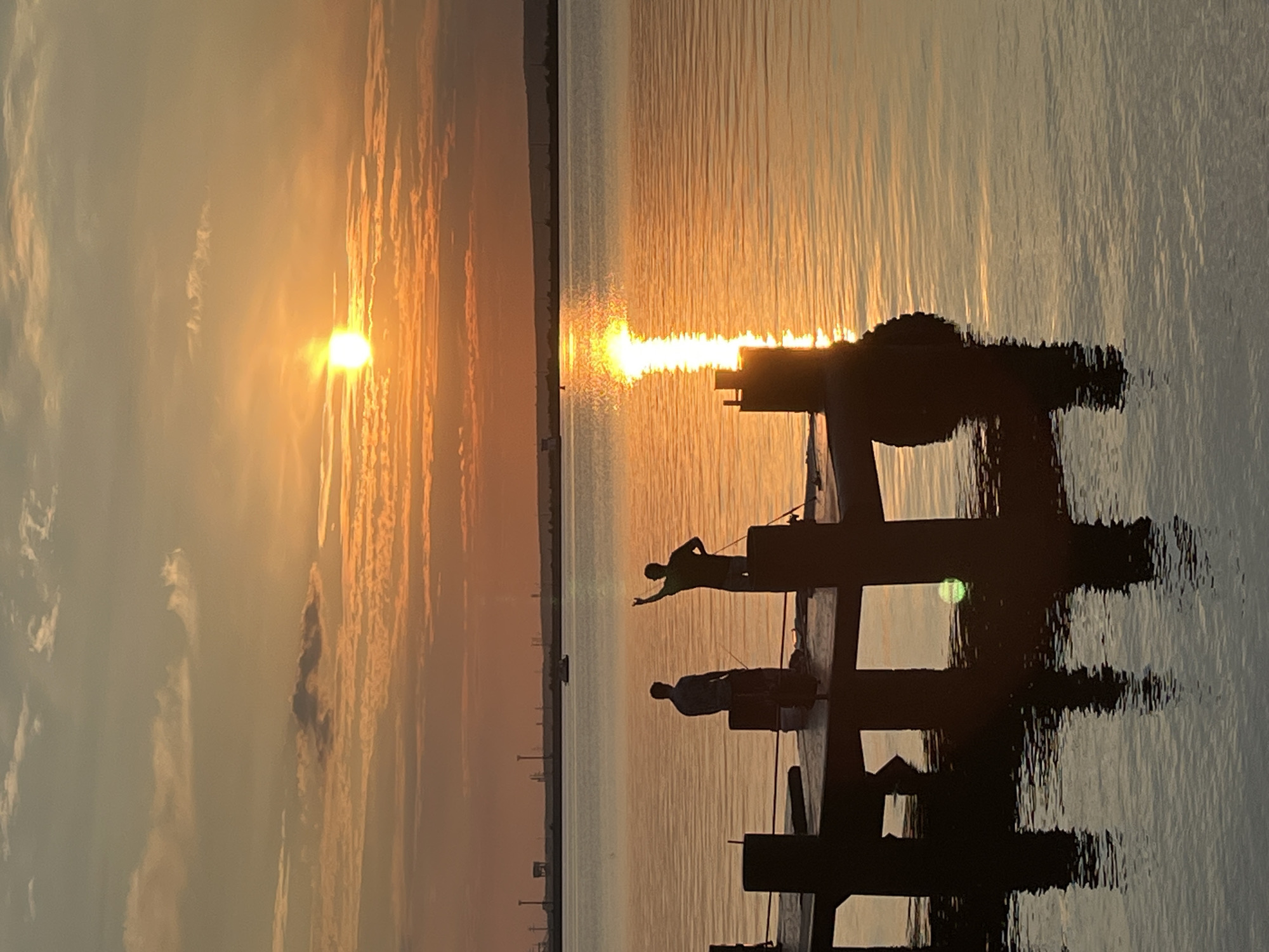 The image depicts a serene sunset over a body of water, likely the Kama River, in Chaikovsky, Russia. Silhouetted figures of two men are visible on a wooden pier. One man is standing with his arm raised, possibly gesturing or casting a fishing line, while the other stands with his back to the viewer. The sun, a bright orb, is setting on the horizon, casting a warm, golden glow across the water and sky, creating a reflective path of light. The sky is filled with soft, streaky clouds, tinged with orange and pink hues from the setting sun. In the distance, the outlines of industrial structures and a small boat can be seen, suggesting the proximity of the city. The water is calm, with gentle ripples reflecting the light and the pier. The overall atmosphere is tranquil and picturesque.