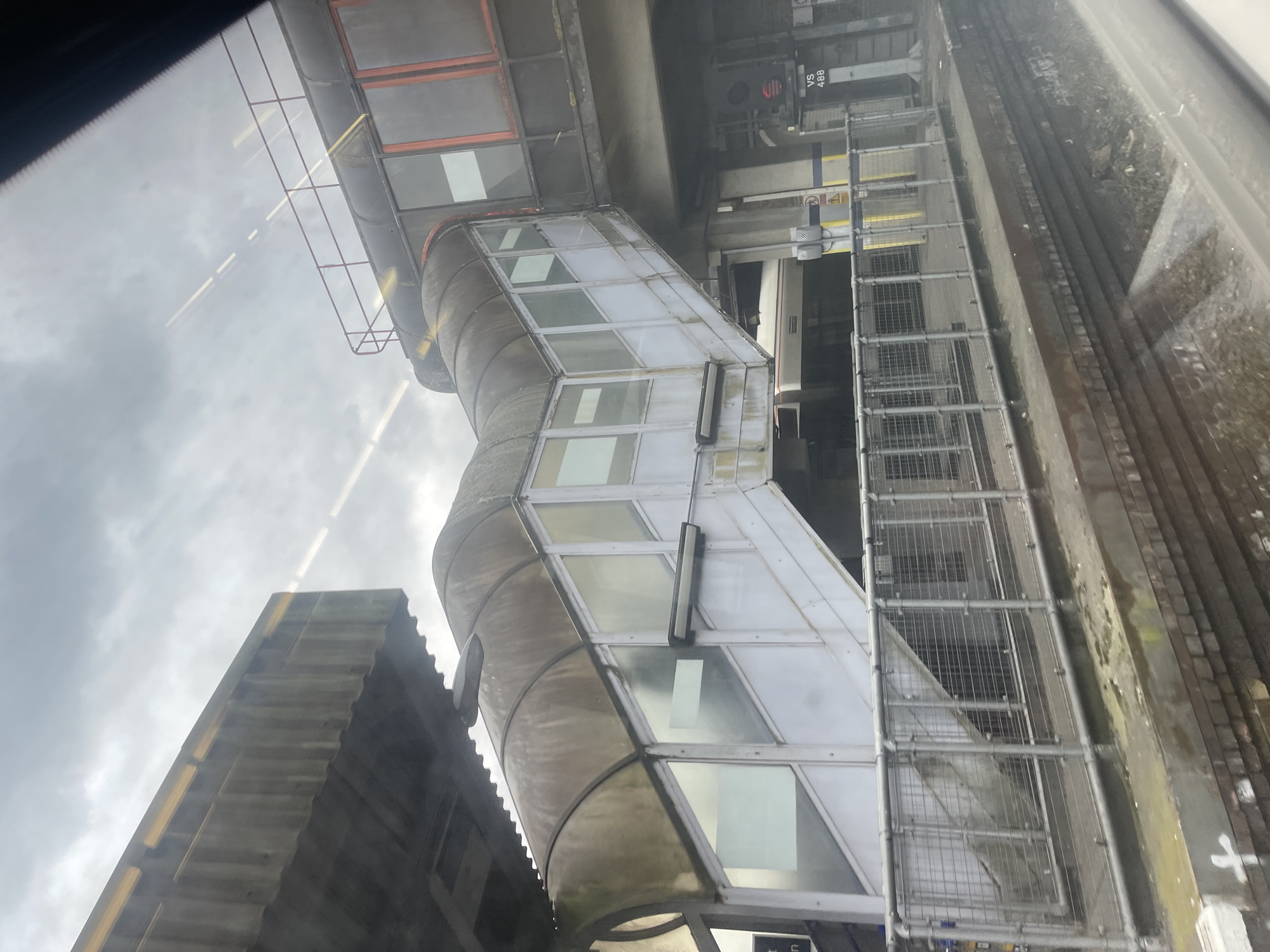 The image depicts a view of Sevenoaks railway station in the United Kingdom. Visible in the shot is the exterior of an elevated passenger walkway connecting different platforms. The walkway is constructed of metal and glass panels. Adjacent to the walkway, there is a metal ladder-like structure running alongside the brick exterior of the rail infrastructure. In the background, there are buildings of the city, and overhead, the sky is overcast.