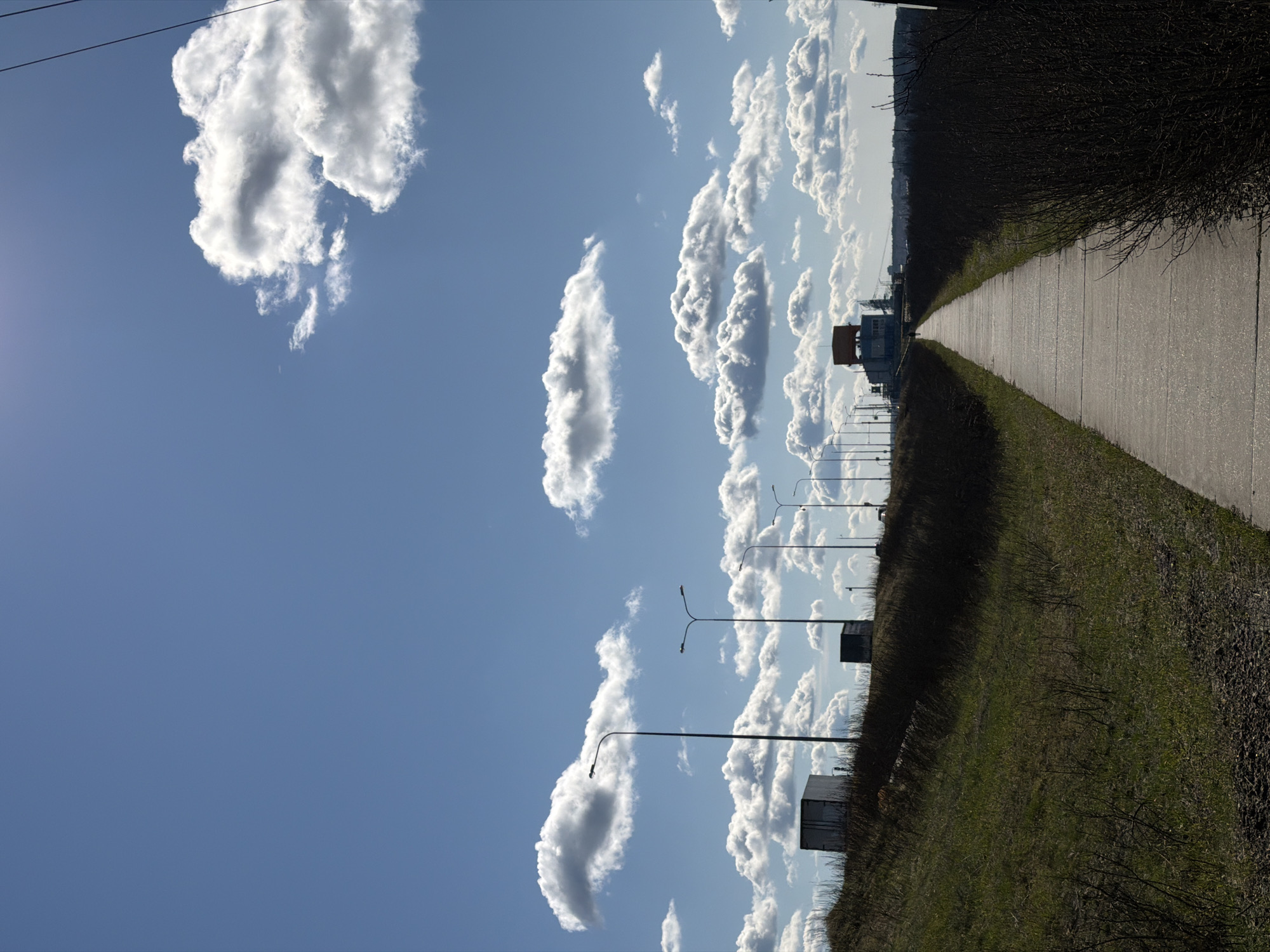 This image, captured in what appears to be a landscape orientation, presents a wide outdoor scene under a clear blue sky filled with numerous fluffy white cumulus clouds. The sun, partially visible in the upper left, casts a bright light, creating a slight lens flare and illuminating the clouds and ground.
In the foreground and extending into the mid-ground on the right side, a paved or concrete path stretches away from the viewer towards the horizon, indicating a long, straight route. This path is bordered by strips of green grass and dense, dark, dry shrubbery or low bushes.
Along the left side of the frame, a series of streetlights with double-arm fixtures recedes into the distance, paralleling the path. In the middle ground on the left, a prominent building with a distinctive orange or yellow roof/top section stands out, suggesting an industrial or utility structure. Further down the path, other smaller, indistinct structures are visible on the left side. Faint overhead wires are also noticeable in the upper left corner.
No people or explicit activities are visible, presenting a calm, expansive view. The bright lighting and distinct shadows indicate it is daytime, likely morning or afternoon. The scene combines natural elements like the sky and vegetation with man-made infrastructure, suggesting an area on the outskirts of an urban or industrial zone, consistent with a location such as Chaikovsky, Russia. There is no visible text in the image.