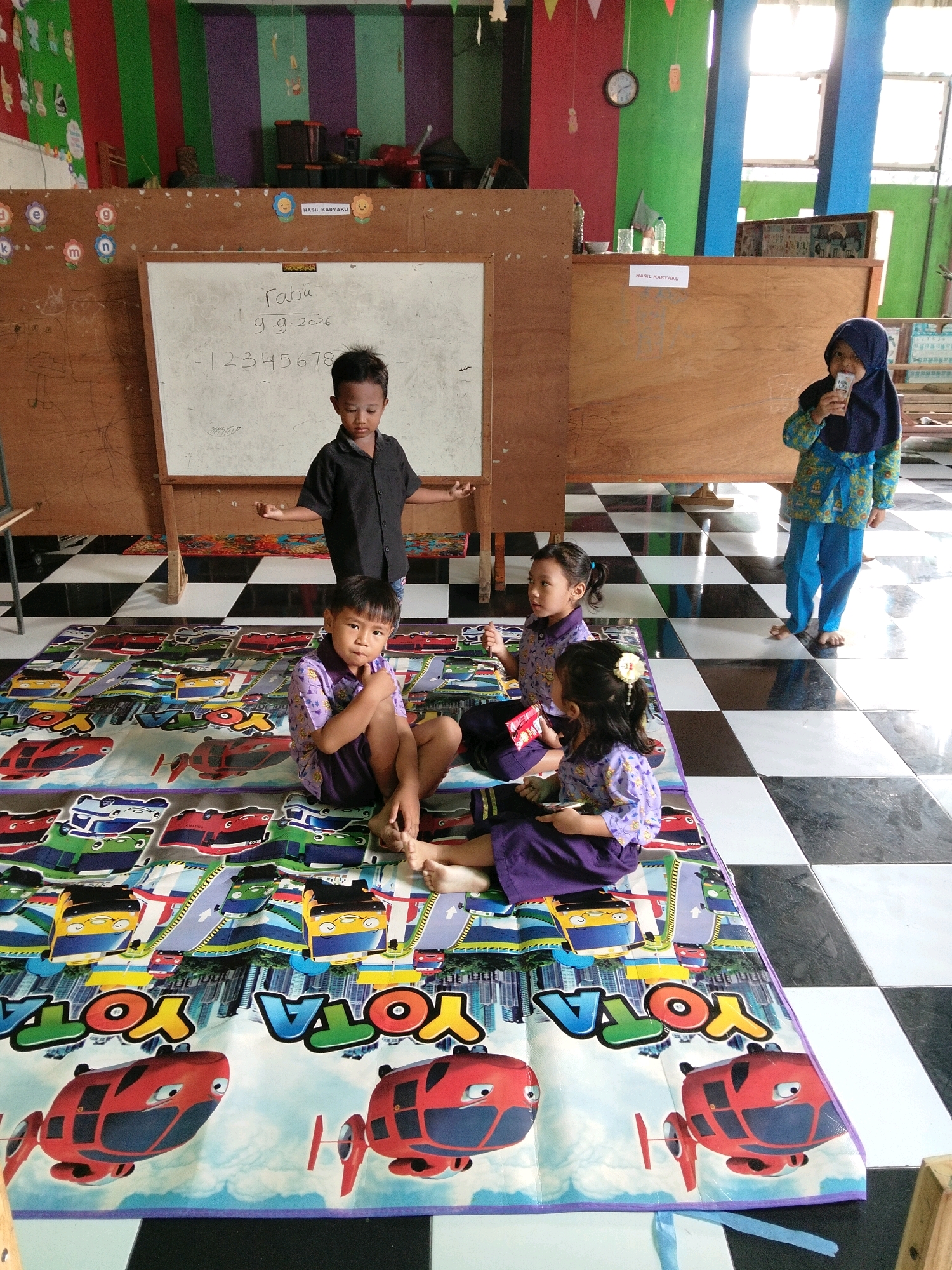 A group of young children are present in a classroom setting, possibly a kindergarten or preschool. One young boy, wearing a black shirt, stands near a whiteboard with handwritten numbers and text. Three other children, two girls and another boy, are seated on a colorful mat depicting cartoon vehicles. One girl is wearing a hijab and holding a small object, possibly a snack. Another girl in a purple outfit has a flower accessory in her hair. The floor is tiled in a black and white checkerboard pattern. In the background, there are colorful walls, wooden partitions, and windows, suggesting an educational environment. The overall scene appears to be a casual indoor activity. The location, Puskesmas Keboan, Indonesia, is a public health center, implying this room might be part of a community center or childcare facility associated with it.