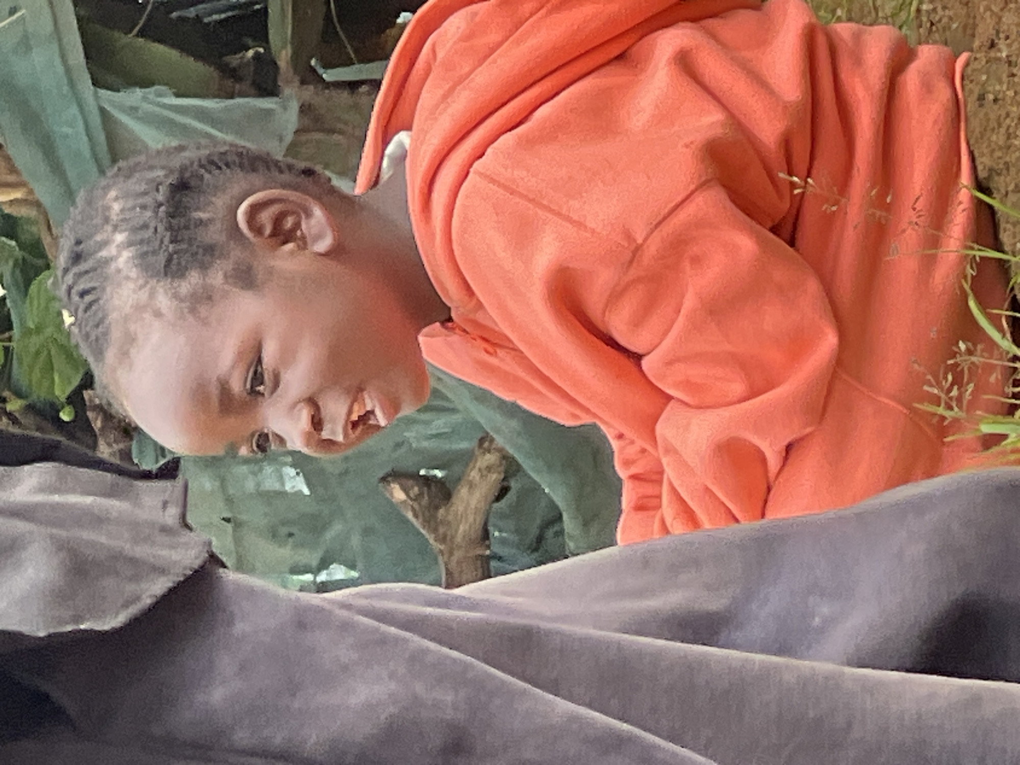 The media file shows a close-up, horizontal shot of a child wearing an orange hoodie. The child's head is tilted to the side, and their gaze is directed downwards and to the left. Their mouth is open, revealing teeth and what appears to be a piece of food or debris. The child's hair is styled in braids.
The setting appears to be outdoors, with hints of greenery and what looks like some sort of tarp or fabric in shades of green and gray. A small, weathered branch is visible behind the child. The overall lighting suggests it is daytime, but no specific time can be determined. The emotions conveyed are difficult to discern due to the angle and lighting, but the open mouth might suggest surprise, exertion, or vocalization. There is no visible text in the image. The context provided places the scene in Jos, Nigeria.