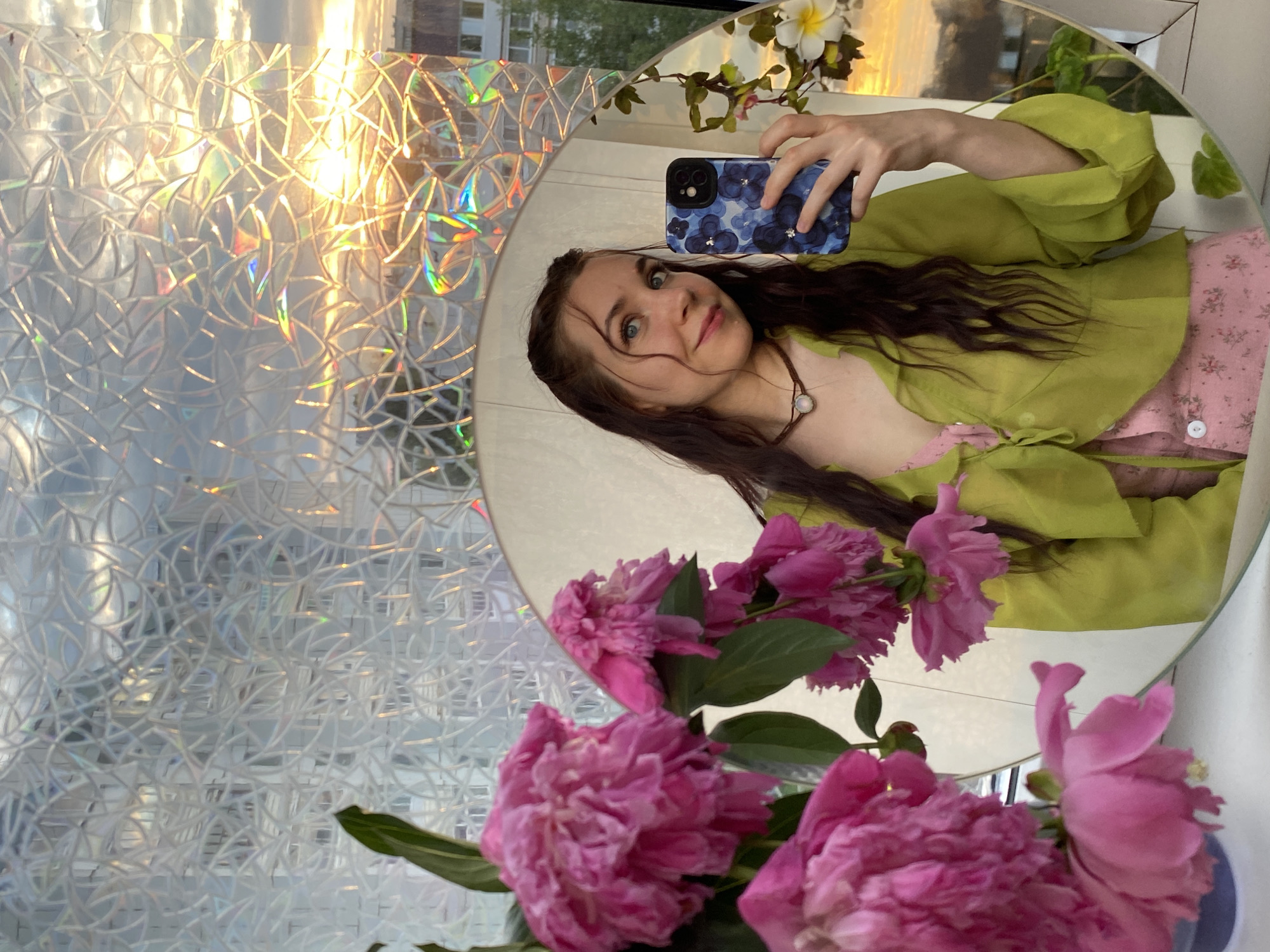 A young woman with long, dark, wavy hair and blue eyes takes a selfie in a round mirror. She is wearing a light green, sheer, button-up blouse over a pink floral dress with small white buttons. A dark brown choker necklace with a single iridescent pendant rests on her neck. She is holding a blue smartphone with a floral pattern in her right hand. The mirror reflects a bouquet of vibrant pink peonies in the foreground and a view of buildings and a sunset through a window with a patterned, textured film. The sunset is bright and casts a warm glow, with visible clouds in the sky. The scene appears to be indoors, likely in an apartment, with the window providing a view of the urban landscape. The background, including the buildings, is slightly out of focus due to the nature of the reflection and the focus on the woman and the flowers. The overall lighting suggests it is either early morning or late evening. This scene is consistent with many apartment balconies or windows in residential areas of Chaikovsky, Russia, particularly during sunset.
