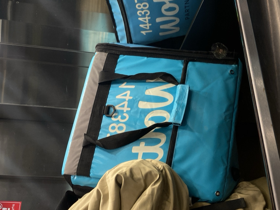 The image displays two blue insulated delivery bags, primarily associated with the Wolt food delivery service, within an interior setting, likely in Tel-Aviv, Israel.
The main subject is a turquoise-blue Wolt delivery bag positioned centrally, with a gray top edge, black fabric straps, and a black zipper. The white lettering "Wolt" is visible in an inverted orientation, partially obscured, along with the numbers "14438". A section of a person's garment, colored light brown or khaki, is visible on the left side, partially covering the bag. The lower right side of this bag exhibits an irregular dark stain or substance near the zipper track. To the right, a second, smaller blue Wolt bag is partially visible, also displaying white lettering "144387 Wolt PARTNE".
The bags rest on a dark surface, with a metallic horizontal bar in the immediate foreground. The background consists of reflective, vertically paneled metallic surfaces. In the upper left corner, a partial red sticker with black Hebrew text, specifically "תק'ן", is visible on the metallic background. No individuals are fully depicted, and no active interactions or specific actions are discernible beyond the placement of the bags. The presence of Hebrew text and the Wolt branding are consistent with operations in Tel-Aviv.