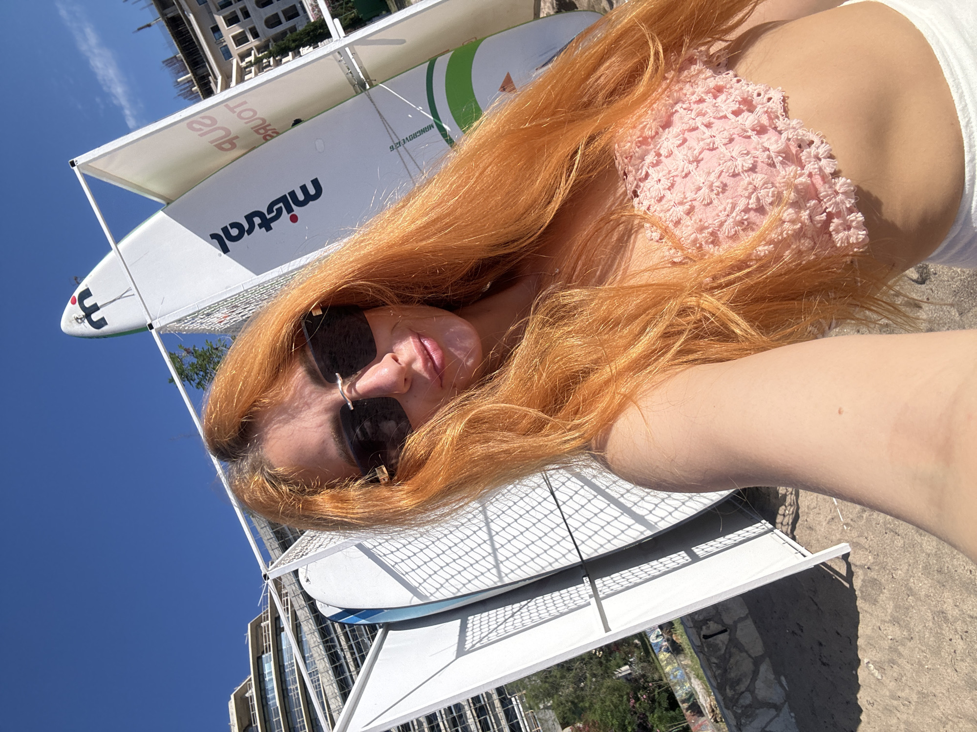 In Budva, Montenegro, on a sunny day, a young woman with long, vibrant orange hair is captured in a selfie. She is wearing large, dark sunglasses and a light pink crochet bandeau top. Her hair is styled loosely, framing her face. In the background, several white paddleboards are propped upright in a rack, some displaying branding such as "Mistral" and "SUP TOURS". Behind the paddleboards, modern buildings with multiple floors and construction scaffolding are visible. The scene appears to be on a beach or boardwalk area, with sand in the foreground and a clear blue sky overhead. The overall impression is a casual, summery atmosphere.