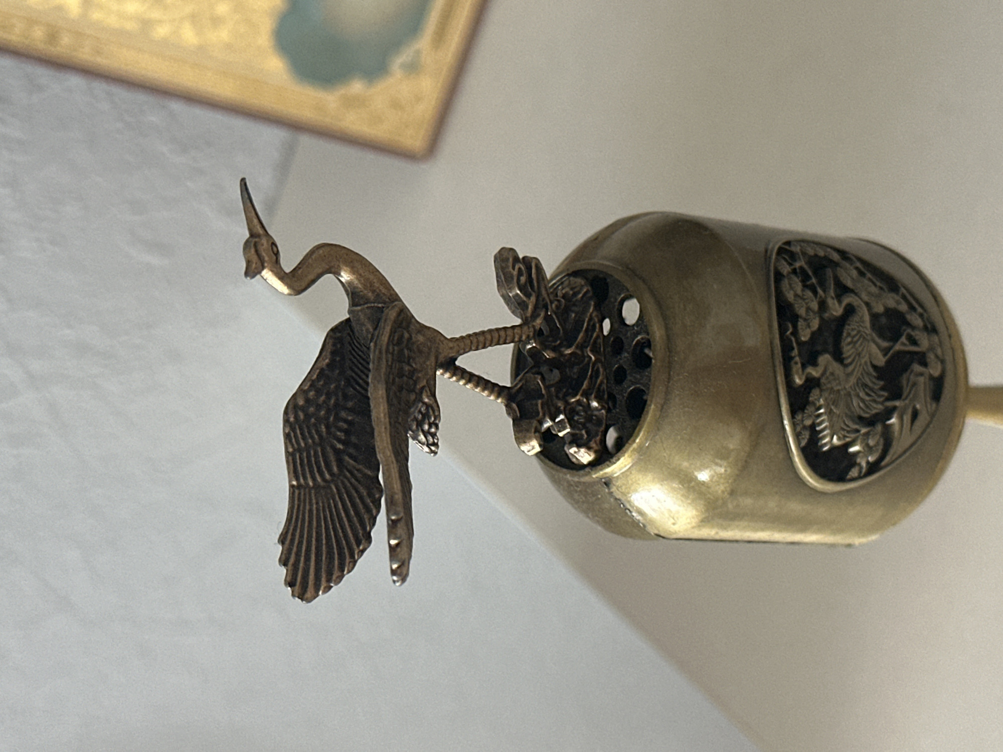 The image shows a close-up, eye-level, slightly low-angle shot of a decorative bronze object with a bird figurine on top. The object is cylindrical and appears to be made of brass or bronze, with intricate carvings of cranes and foliage on its surface. The bird figurine, also bronze, is depicted with wings outstretched and standing on what looks like a stylized floral or rocky base. The background is a plain, light-colored surface, possibly a wall or fabric, with a hint of a golden frame visible in the upper right corner, suggesting the object might be part of a larger display or collection. The lighting is soft, casting subtle shadows. The image does not contain any information or context related to the city of Nevinnomyssk, Russia.