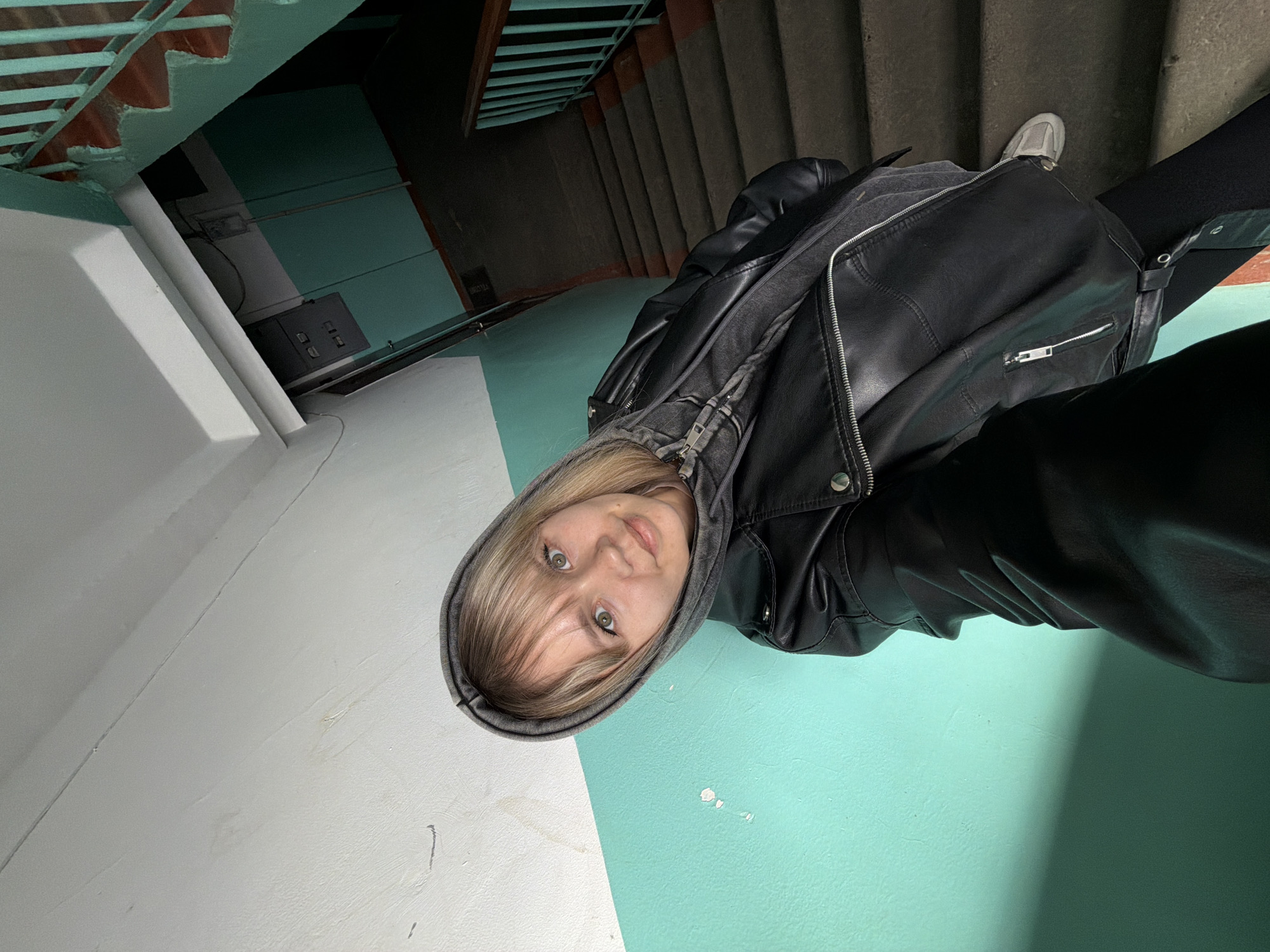 A young woman is depicted in a rotated selfie, looking directly at the camera with a neutral to slightly curious expression. She has blonde hair with bangs, green eyes, and is wearing a grey hooded sweatshirt underneath a black leather jacket with silver zippers and hardware. Her left shoe, a light grey sneaker, is visible in the upper right corner of the frame, resting on a stair.
The setting appears to be an indoor stairwell or hallway, characterized by concrete stairs and dark metal railings visible in the background. The walls are painted in two distinct sections: a bright white on the left and a prominent teal or turquoise on the right, extending into the background. A white pipe runs vertically along the left wall. Further back on the teal wall, a light fixture and a grey electrical box with several controls are visible. The lighting is somewhat dim, suggesting an indoor environment, possibly with artificial light or limited natural light.
The woman is the central focus, positioned against the dividing line of the two wall colors. The image captures a casual moment, likely a self-portrait taken in a public or communal space within a building. No discernable text is visible anywhere in the image.