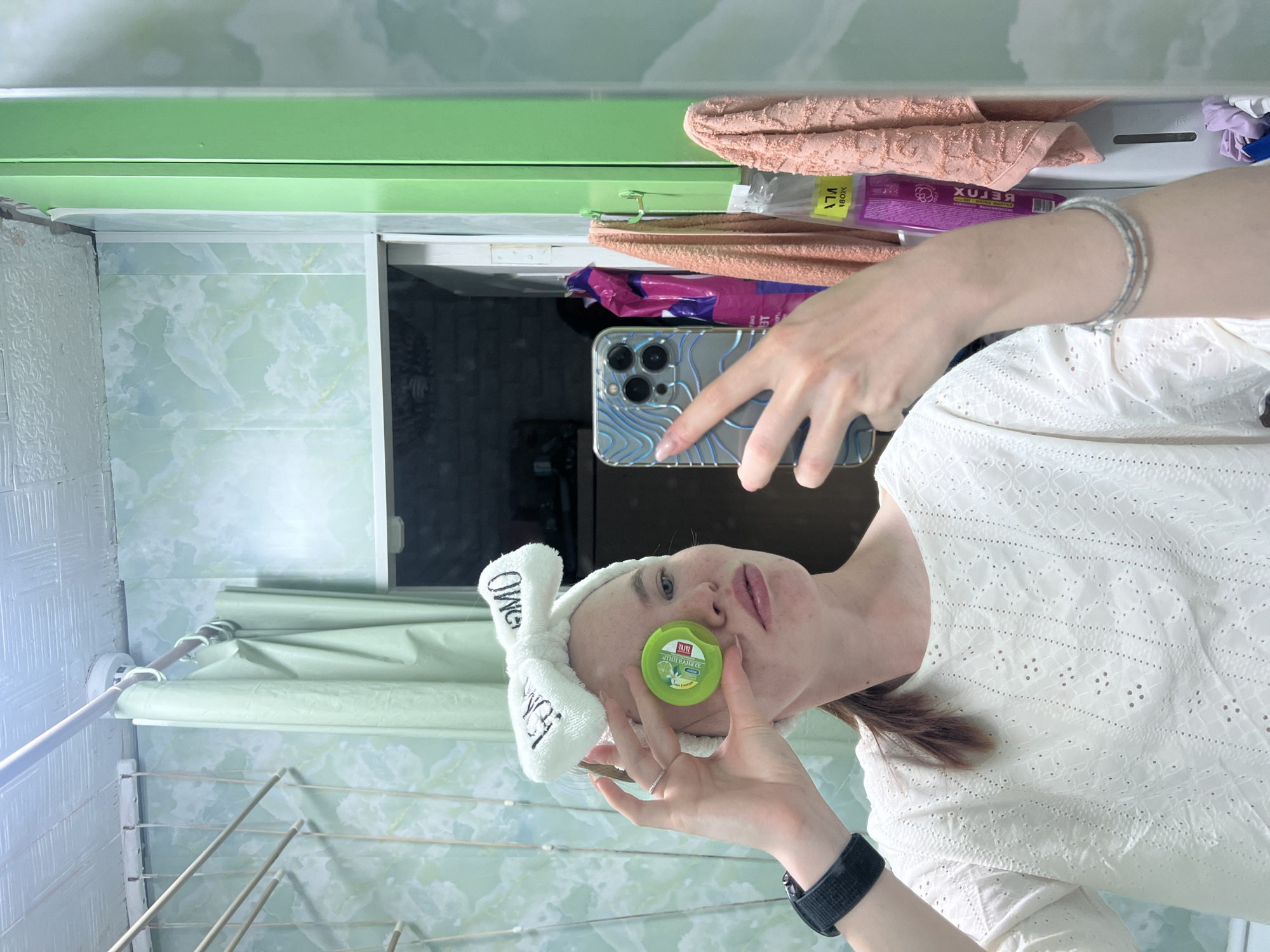 A young, light-skinned woman stands in a bathroom with green and white marble-patterned walls. She wears a white head towel with the text "OMG" and a white eyelet top. She holds a small, round, green container with text in front of her face. Her left hand holds a smartphone with a blue and silver patterned case as she takes the photograph. A black smartwatch is on her left wrist. Above her, there is a green cabinet with towels and other items stored on top. Below, there is a clothes drying rack and a shower curtain. The location is Chaikovsky, Russia, as indicated by the Cyrillic text on the green container.