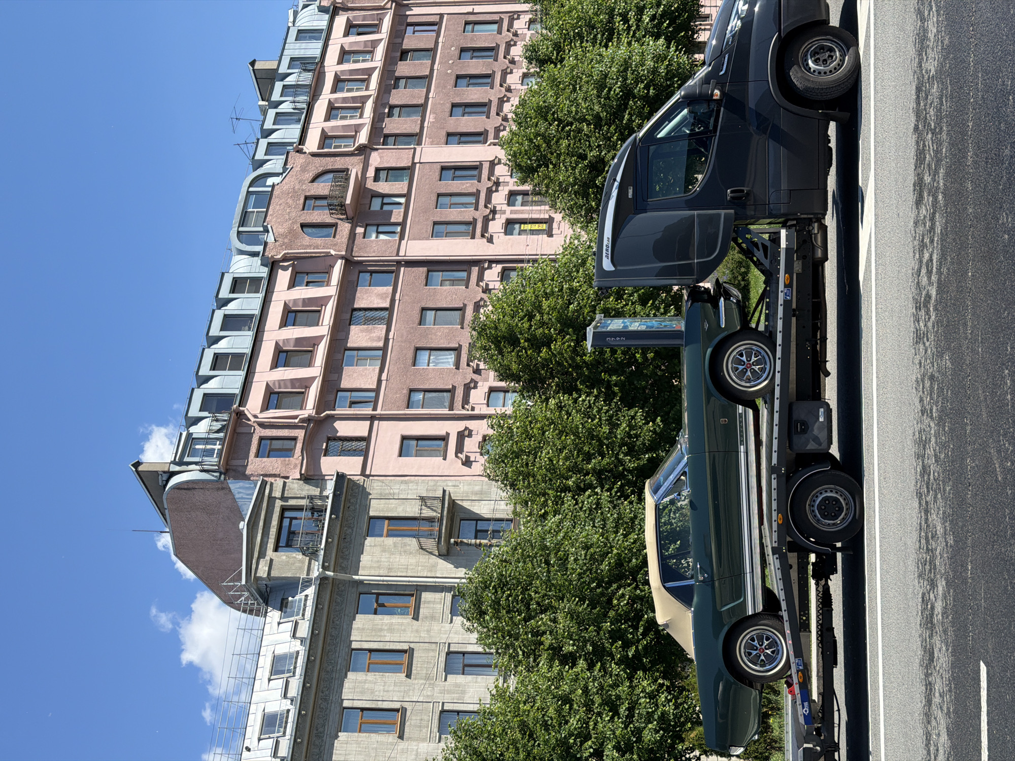 A dark grey flatbed tow truck is parked on a paved road in Saint Petersburg, Russia, under a clear blue sky. The tow truck is transporting a vintage dark green convertible classic car, appearing to be a Ford Mustang. The car is positioned on the flatbed with its wheels secured. Behind the tow truck, a line of lush green trees forms a dense canopy. Beyond the trees, a row of multi-story buildings with pink and grey facades are visible. The buildings have numerous windows and ornate architectural details, typical of Saint Petersburg's historic cityscape. The scene appears to be captured during daylight on a sunny day.