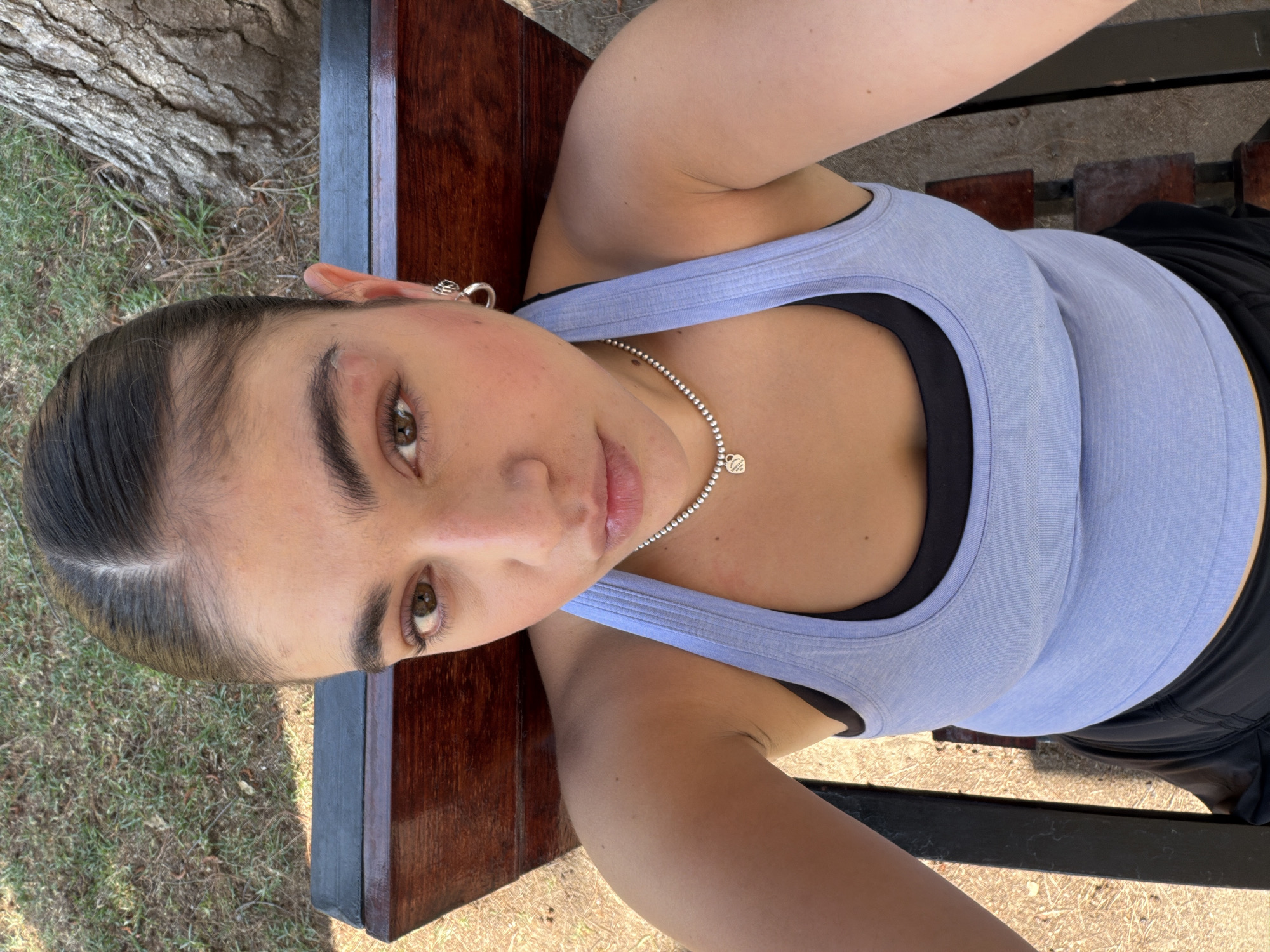 A woman is lying on a wooden bench outdoors. She has dark hair pulled back, brown eyes, and is wearing a light blue tank top over a black sports bra, with a silver necklace. Her head is near the edge of the bench, which has a dark-stained wooden surface and a black metal frame. The ground around the bench is covered in grass and patches of dry earth. In the background is a tree. There is no way to definitively identify the location as Puebla, Mexico based solely on the visual information provided in the image.